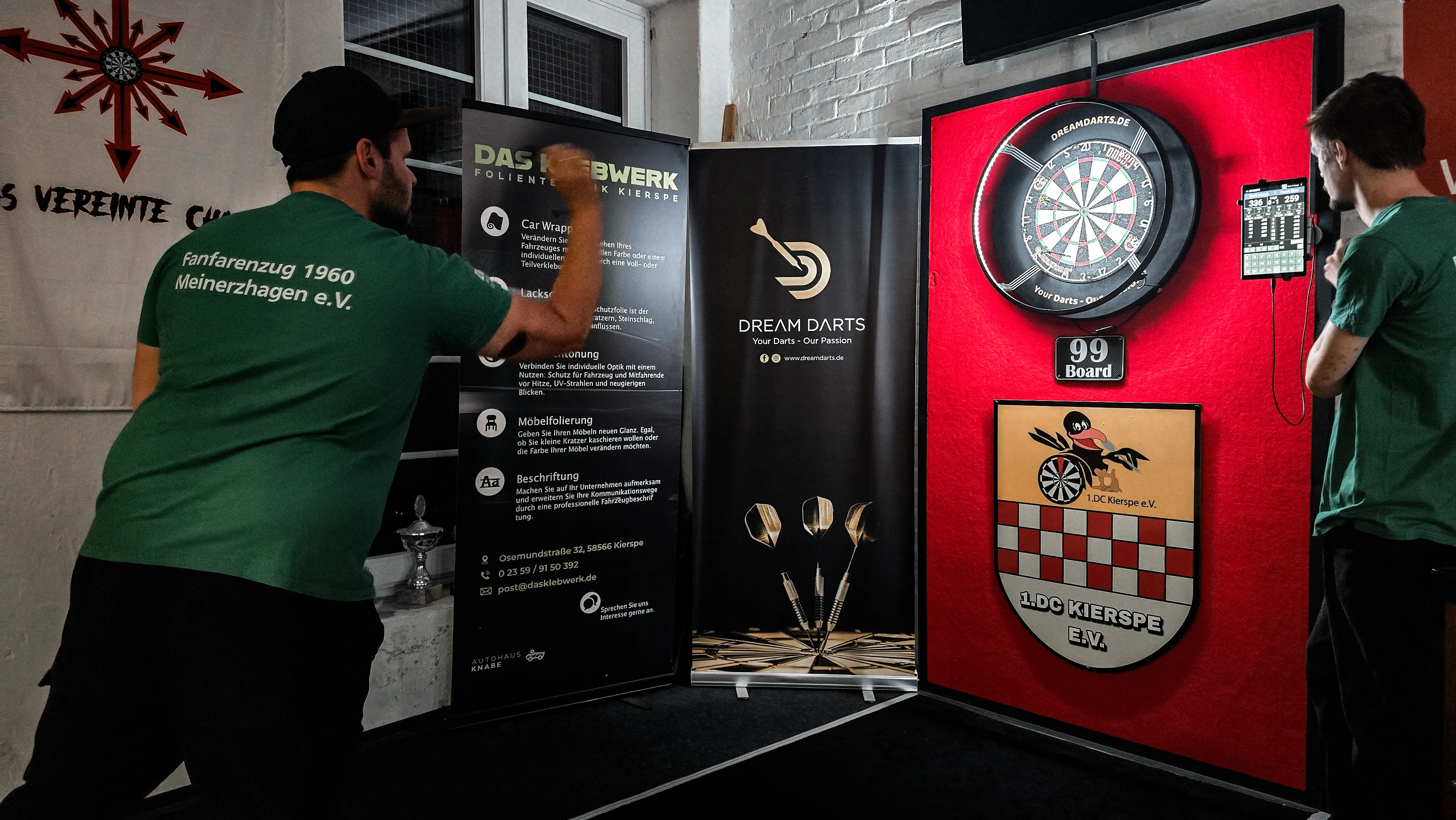 Dartscheibe an einer Wand mit Werbeaufdrucken für Folientechnik und den Dartclub Kierspe, im Vordergrund ein Pokal und ein Wappen des Fanfarenzugs Meinerzhagen.