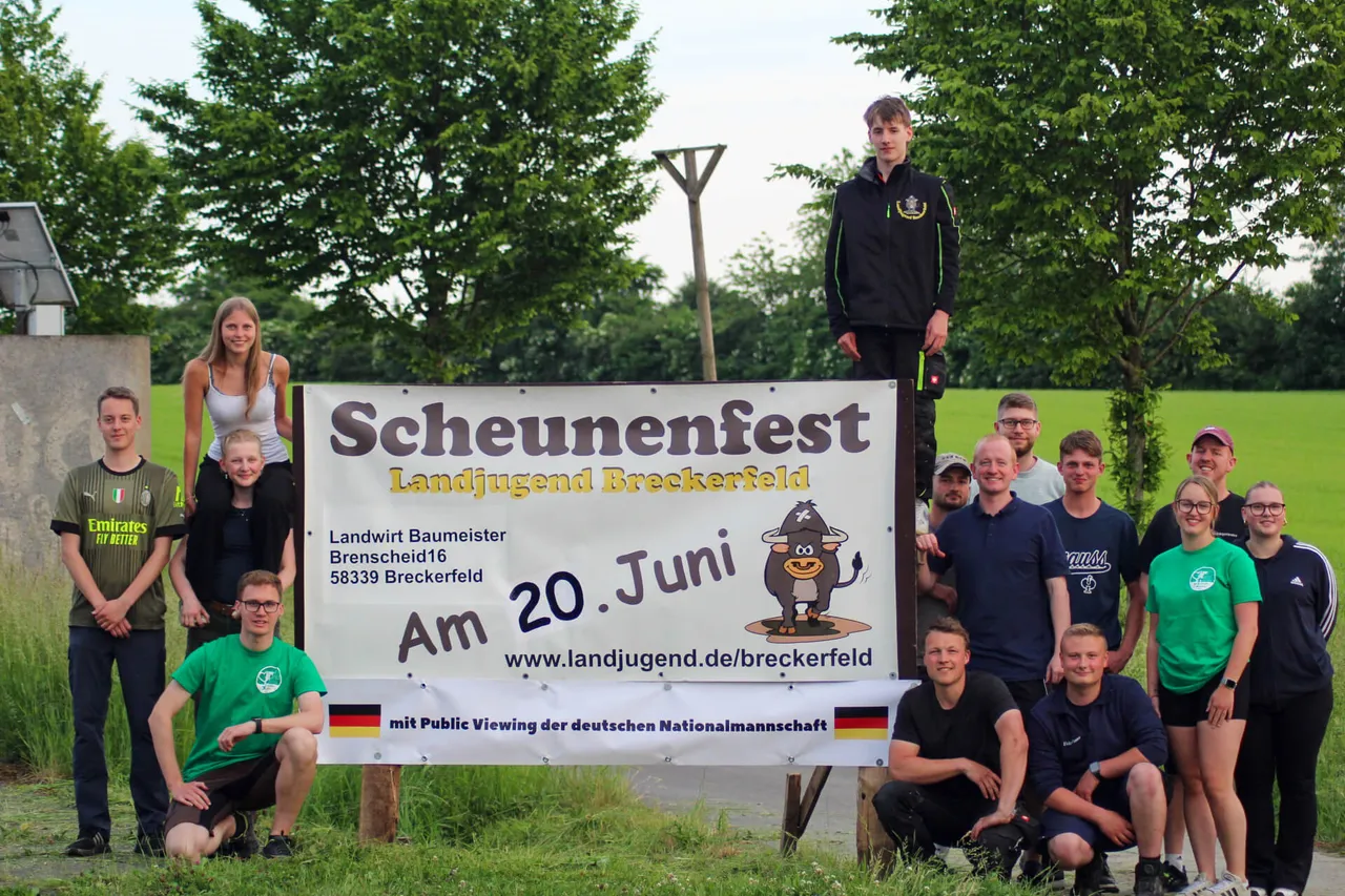 Gruppe junger Menschen steht vor einem Banner mit der Aufschrift „Scheunenfest“ und dem Datum 20. Juni. Im Hintergrund sind Bäume und ein Fußballtor zu sehen.