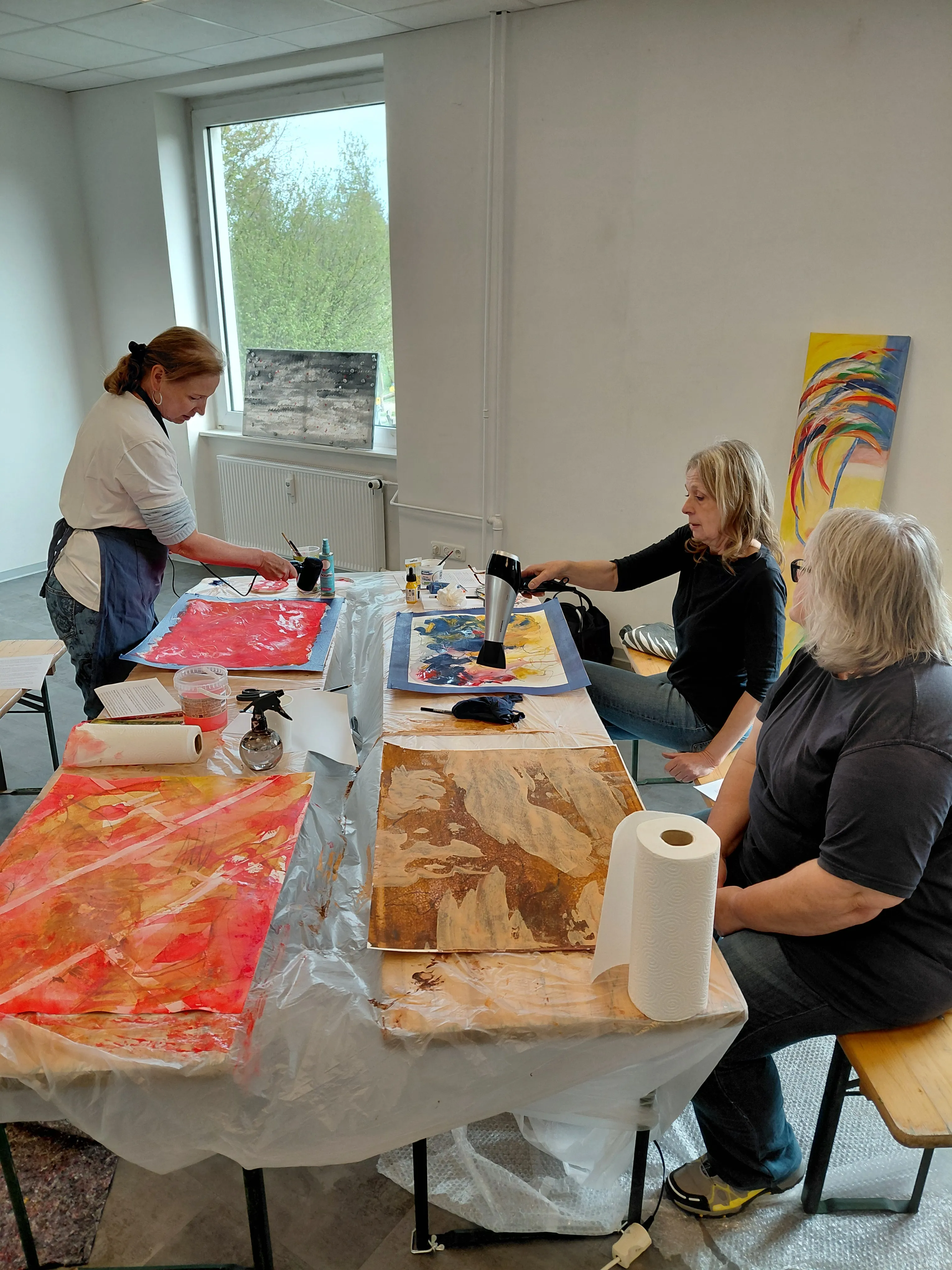 Drei Frauen sitzen an einem Tisch und arbeiten an abstrakten Gemälden mit Acrylfarben. Eine Frau hält einen Fön, um die Farbe zu trocknen.