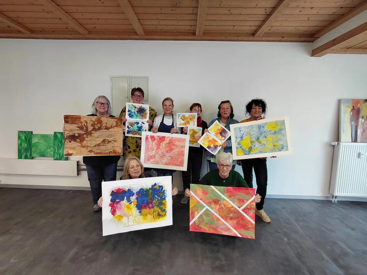 Eine Gruppe von sieben Frauen hält ihre selbstgemalten abstrakten Bilder in einem hellen Atelier vor. Die Bilder sind in verschiedenen Farben und Stilen gehalten.