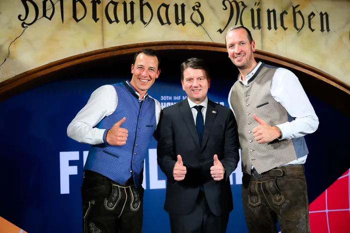 Drei Männer posieren vor einem Hintergrund mit dem Schriftzug „Hofbräuhaus München“ und dem Logo der Handball-Weltmeisterschaft. Von links: Dominik Klein, Sandro Kirchner und Pascal Hens geben alle Daumen hoch.