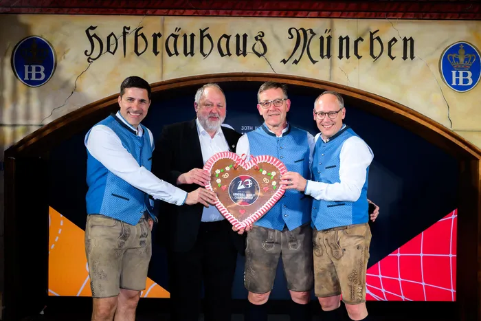 Vier Männer in Tracht und Anzug stehen im Hofbräuhaus München und halten eine runde, rot-weiß verzierte Trophäe mit der Aufschrift „20 Hensoldt World Championship“ in den Händen.