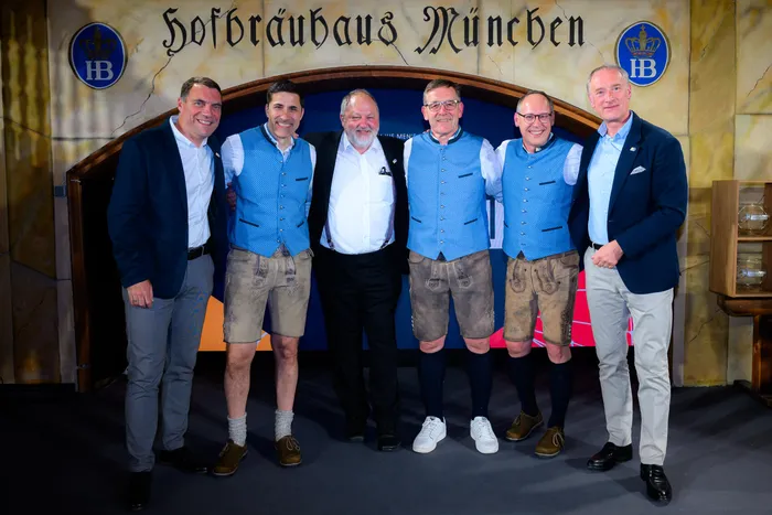 Fünf Männer in Trachten stehen vor dem Eingang des Hofbräuhauses in München. Sie tragen karierte Hemden, Lederhosen und Jacken, einige mit Ansteckern versehen.