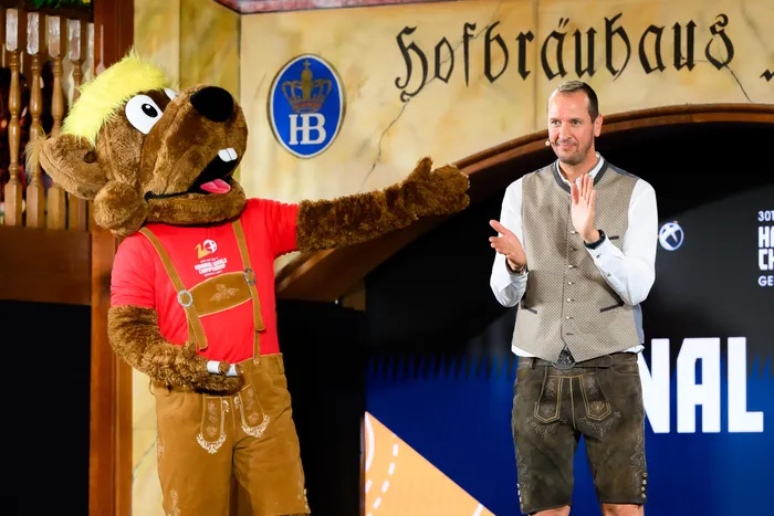 Ein Mann in Tracht und ein Maskottchen in rot-weißem Trachtenhemd stehen nebeneinander und lächeln in die Kamera. Im Hintergrund ist ein Schild mit der Aufschrift „Hofbräuhaus“ zu sehen.