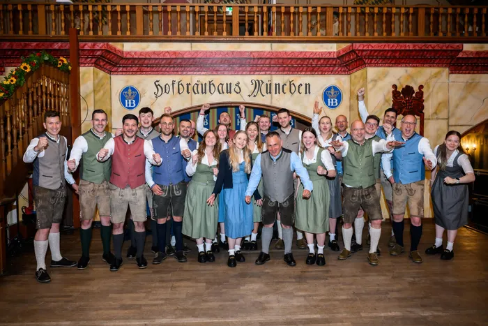 Eine Gruppe von etwa 20 Personen in Tracht steht vor einer Wand mit dem Schriftzug 