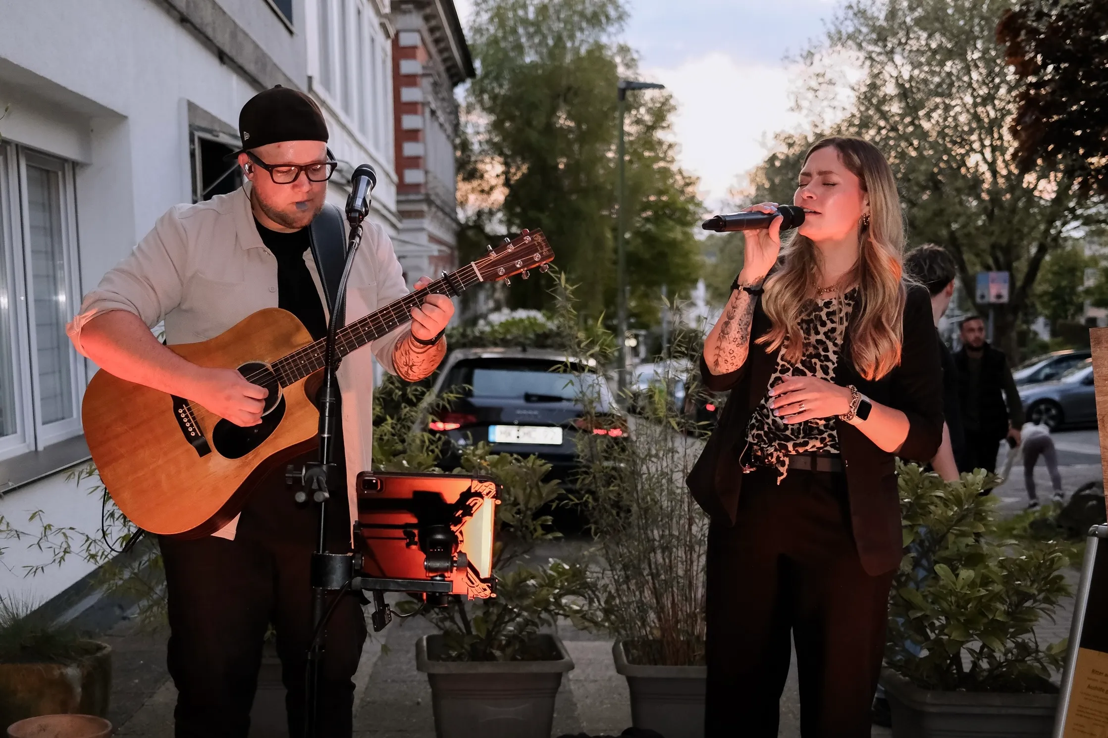 Ein Mann spielt Gitarre und eine Frau singt in ein Mikrofon auf einer Straße, während im Hintergrund Menschen vorbeigehen.