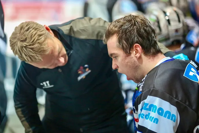 Mannschaftsarzt Dr. Jochen Veit beugt sich zu Eishockeyspieler Sven Ziegler auf der Spielerbank vor.