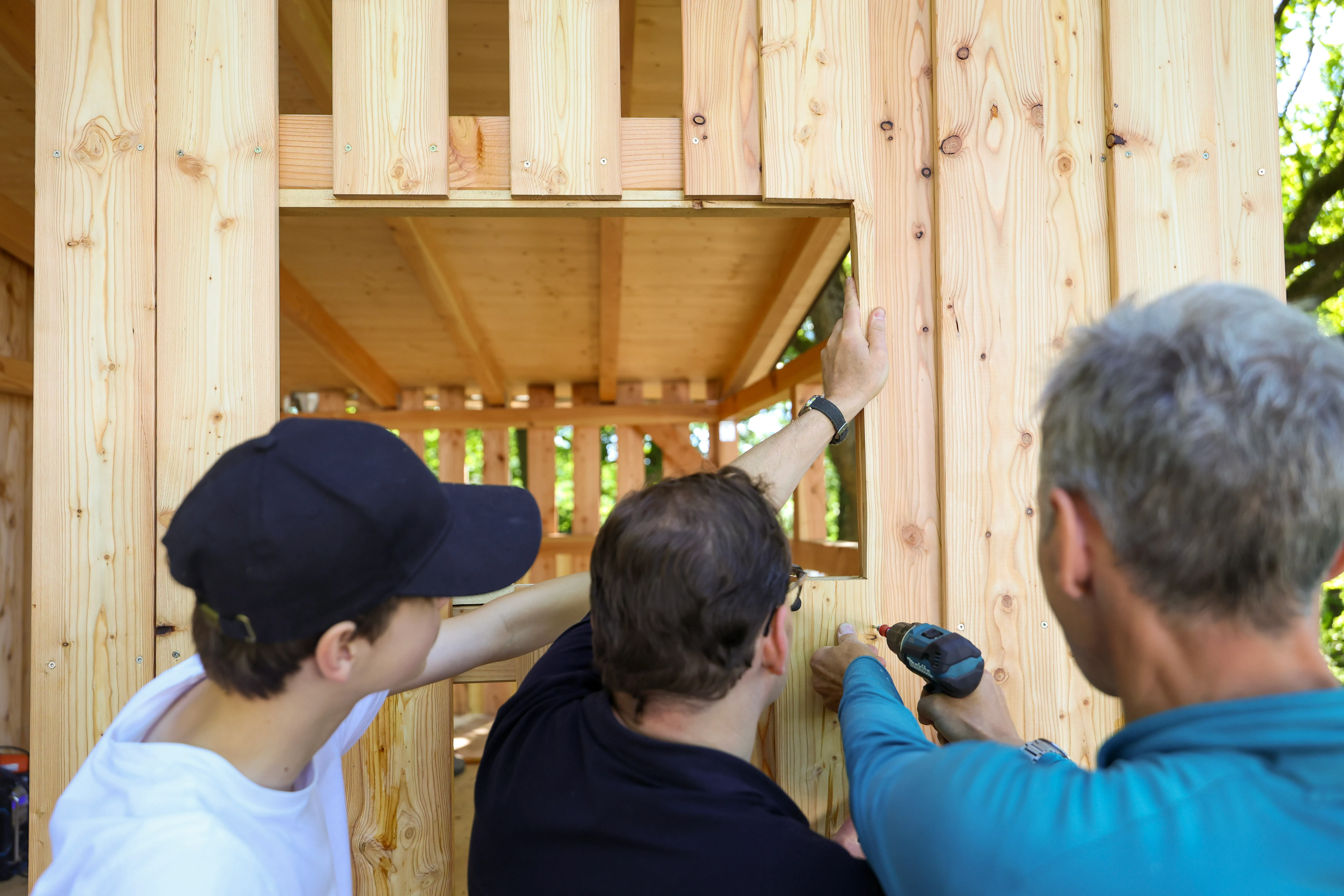 Drei Personen bauen eine Holzkonstruktion, vermutlich eine Hütte, mit Werkzeug. Im Hintergrund ist die weitere Holzverkleidung der Hütte zu sehen.