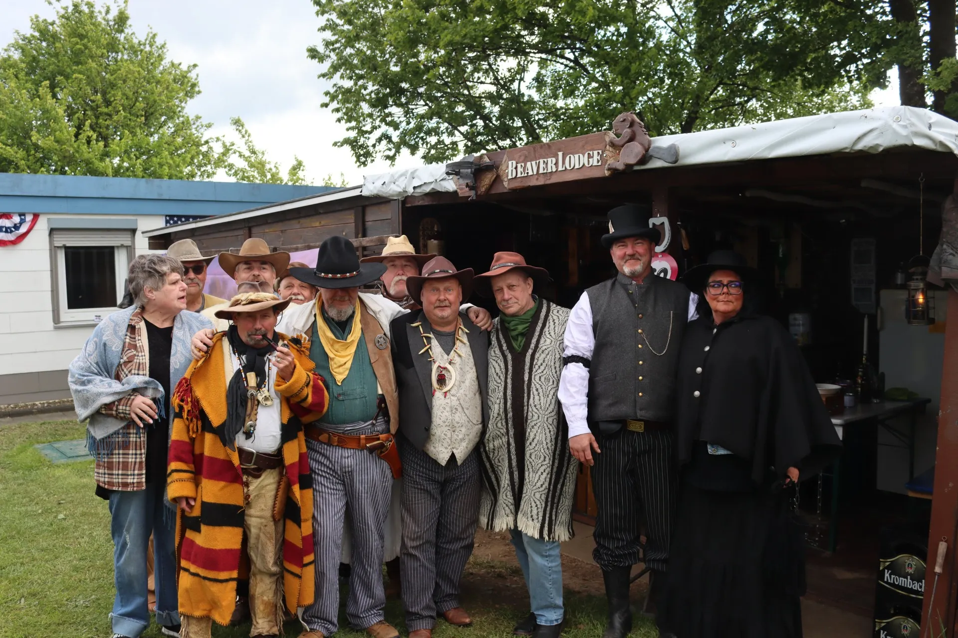 Eine Gruppe von Menschen in Western-Kostümen steht vor einem Holzgebäude mit dem Schriftzug „Beaver Lodge“. Sie tragen Hüte, Westen und karierte Hemden und posieren für ein Foto.