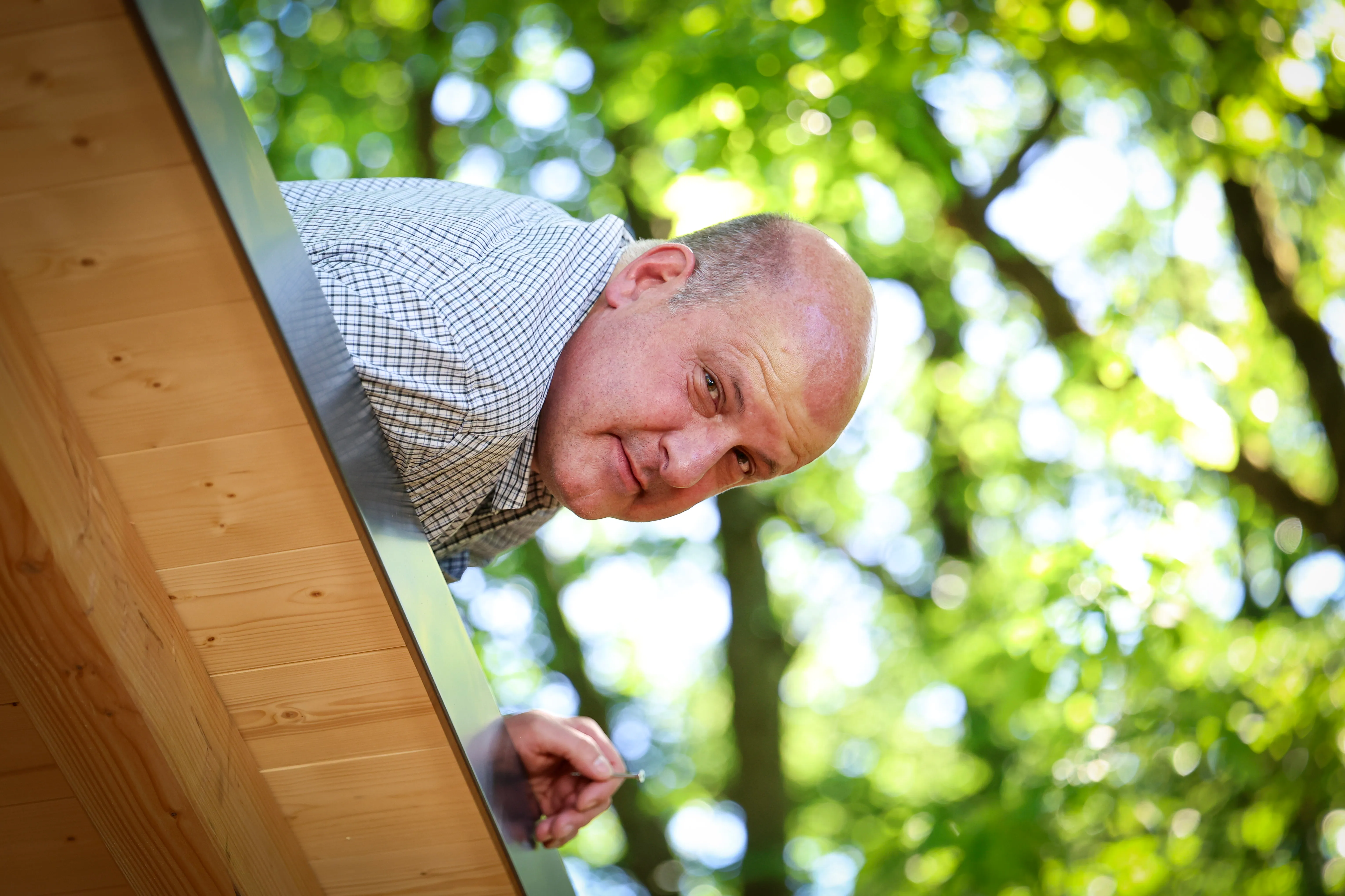 Ein Mann mit Glatze und kariertem Hemd lächelt über eine Holzkonstruktion, im Hintergrund grünes Laub.