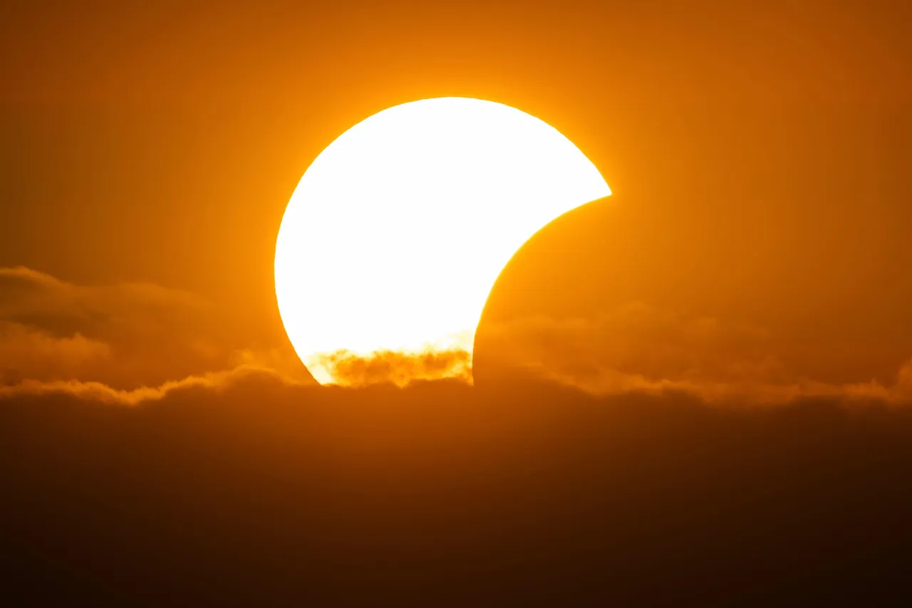 Eine helle, sichelförmige Sonne während einer partiellen Sonnenfinsternis über einer Wolkendecke vor einem orangefarbenen Himmel.