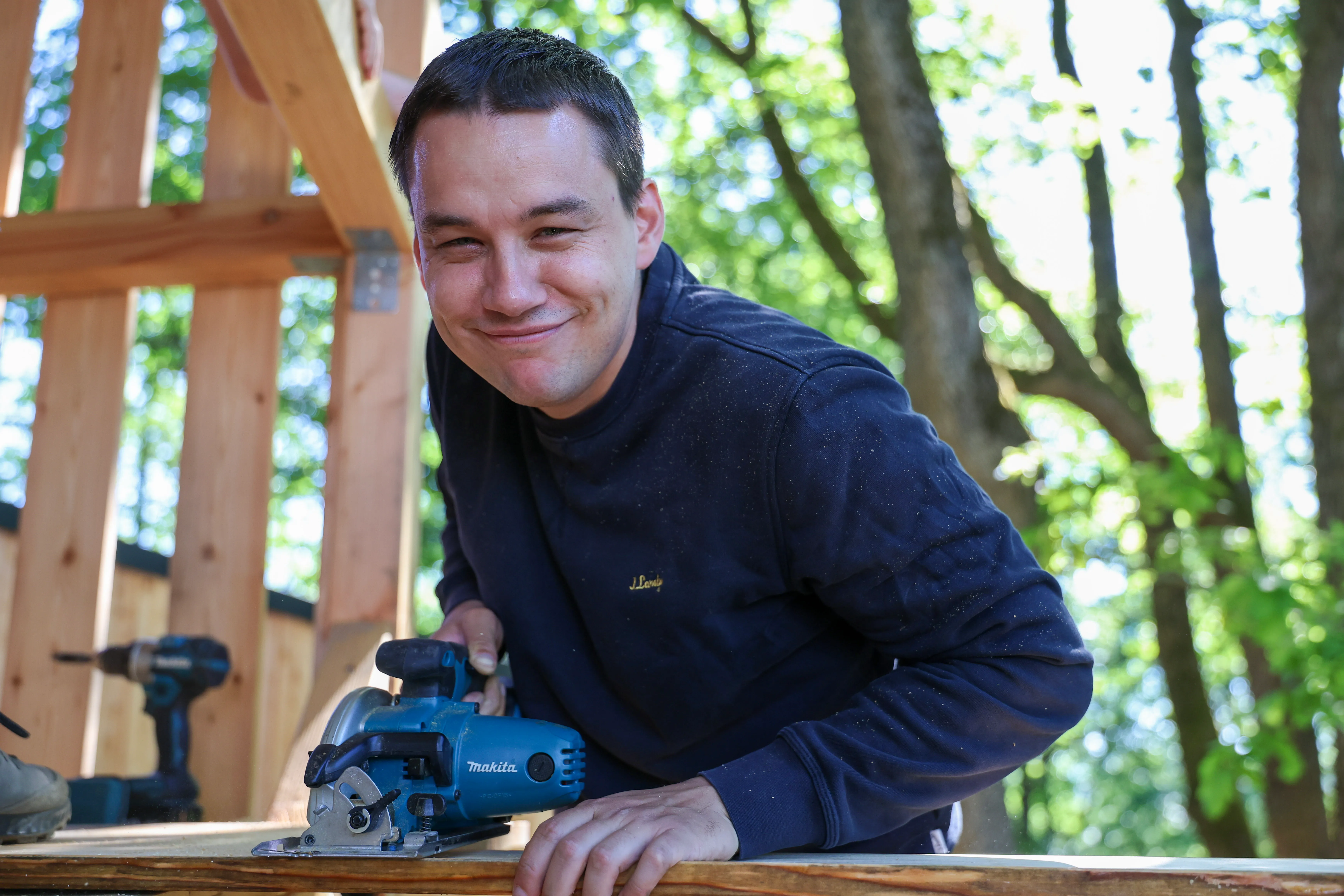 Ein Mann lächelt in die Kamera, während er eine Kreissäge auf einem Holzbrett bedient. Im Hintergrund sind Holzbalken und grüne Bäume zu sehen.