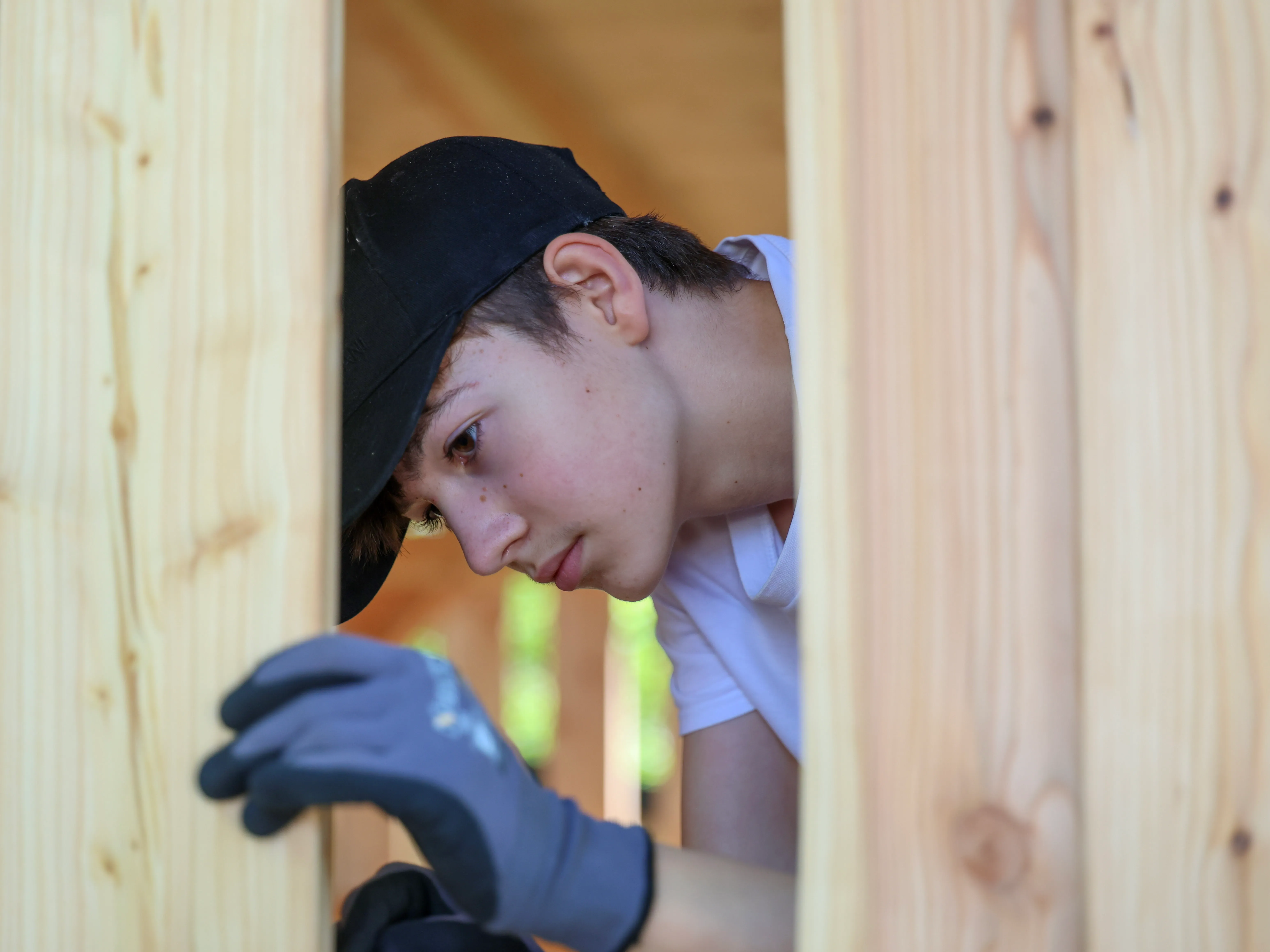 Ein Jugendlicher mit Baseballkappe und Arbeitshandschuhen blickt konzentriert durch eine Öffnung in einer Holzwand.