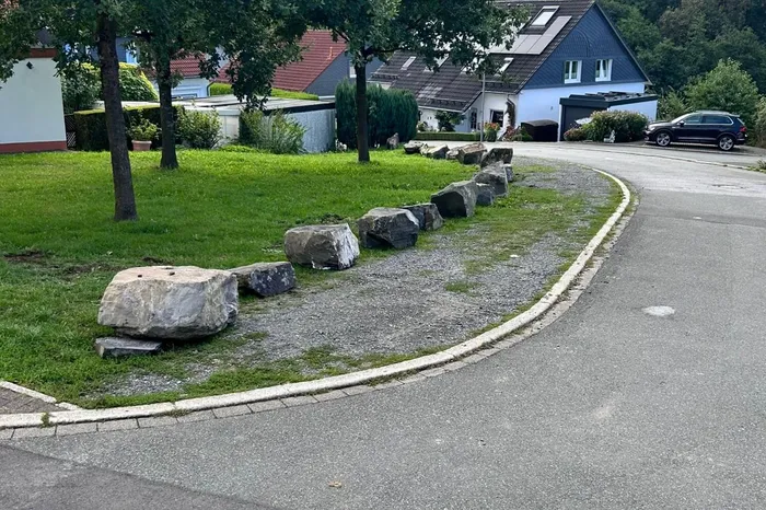 Eine Reihe großer Findlinge säumt den Rand einer Rasenfläche neben einer asphaltierten Straße in einem Wohngebiet.