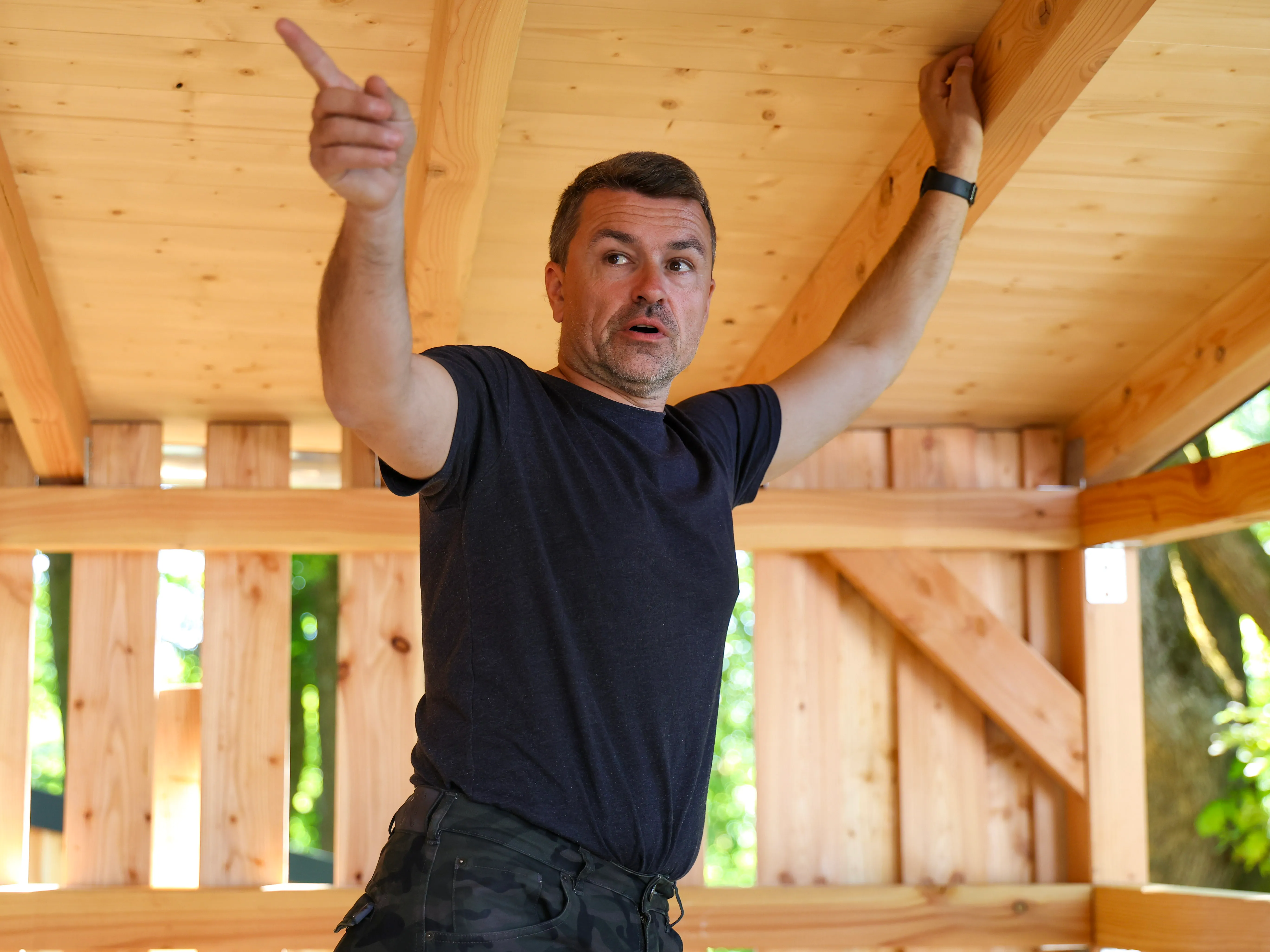 Ein Mann in Arbeitskleidung zeigt mit ausgestrecktem Arm nach oben in den Rohbau einer Holzkonstruktion.