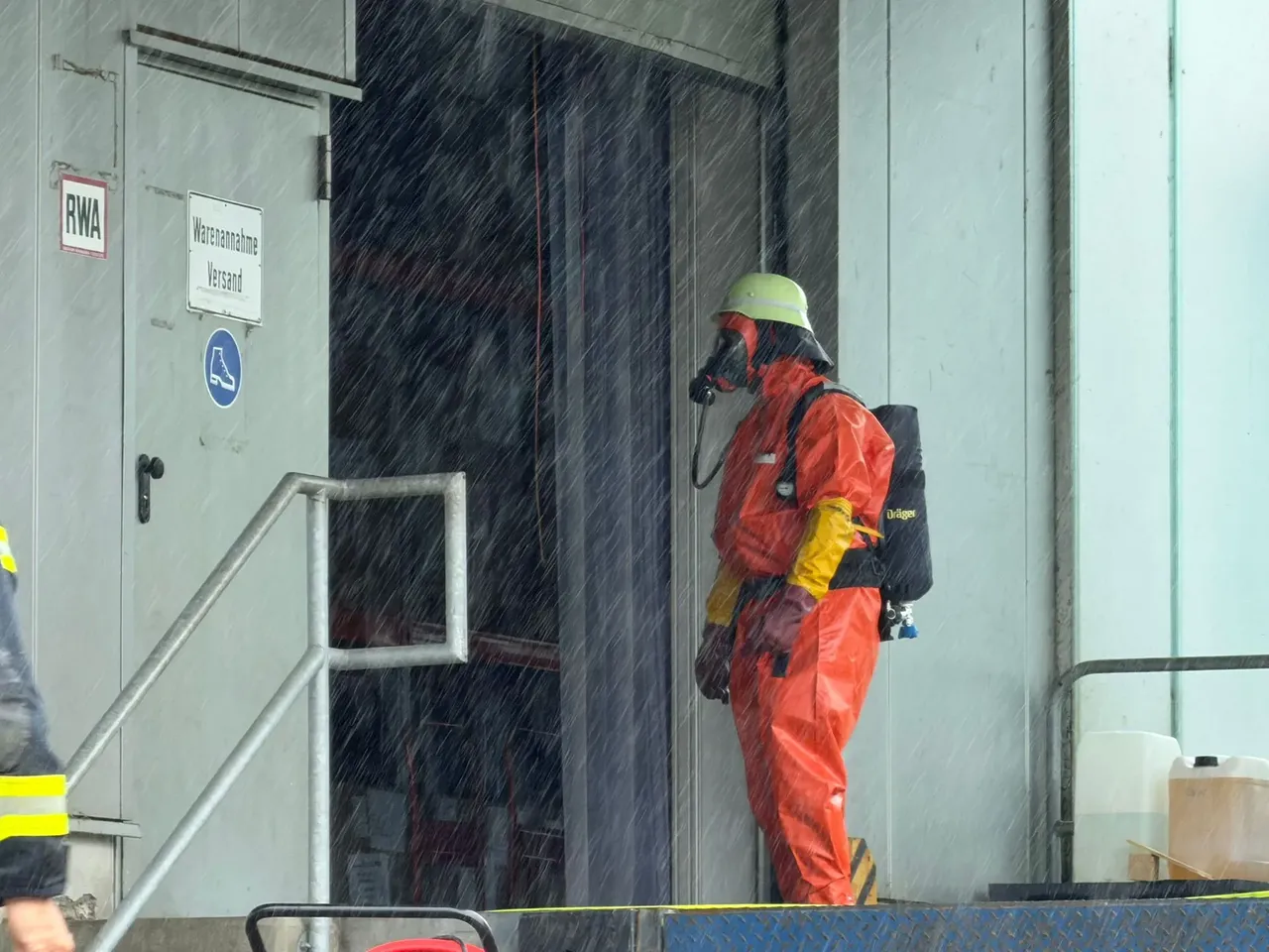Ein Feuerwehrmann in einem Schutzanzug steht im strömenden Regen vor einem Gebäude. Er trägt eine Atemschutzmaske und einen Sauerstofftank auf dem Rücken. Ein Schild über der Tür weist auf 'Warenannahme' und 'Versand' hin.