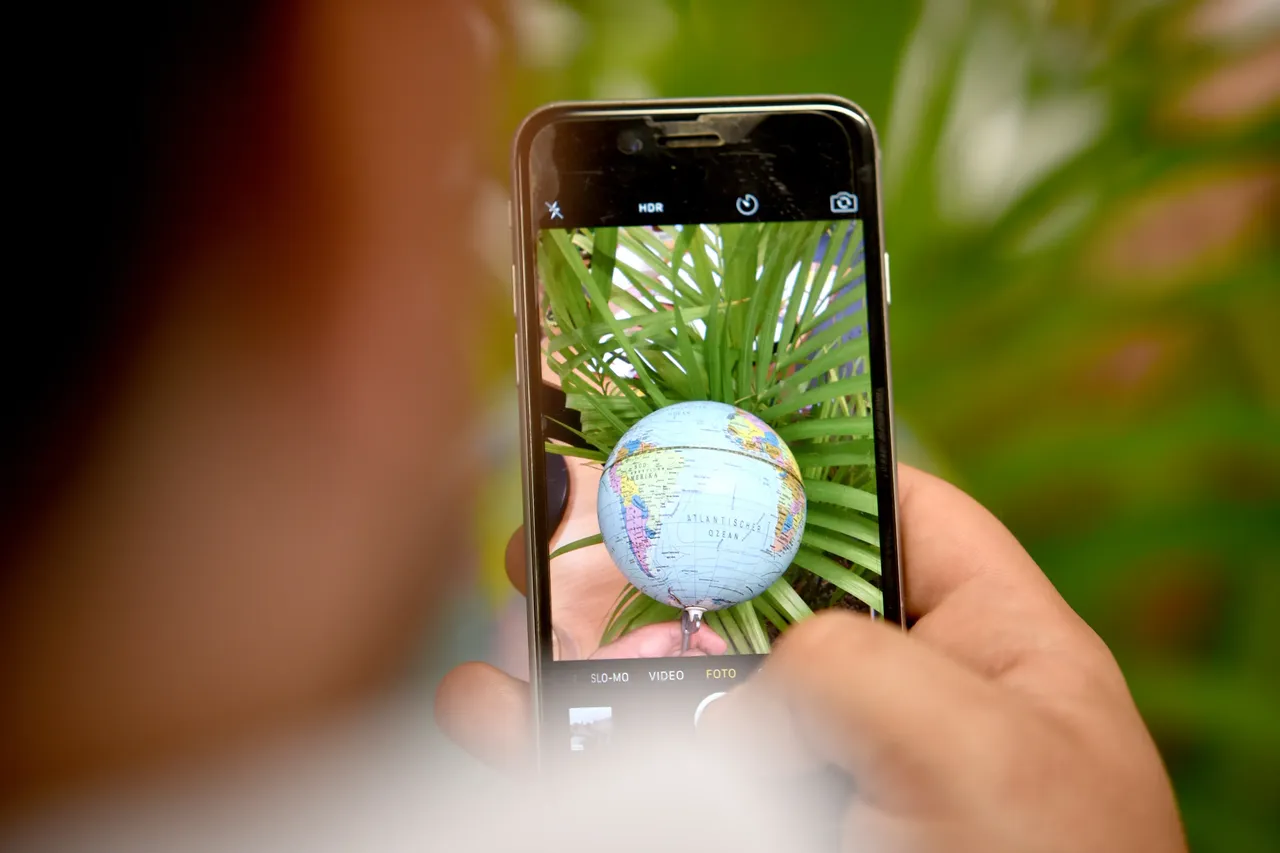 Eine Person fotografiert mit einem Smartphone einen kleinen Globus, der zwischen grünen Blättern liegt.