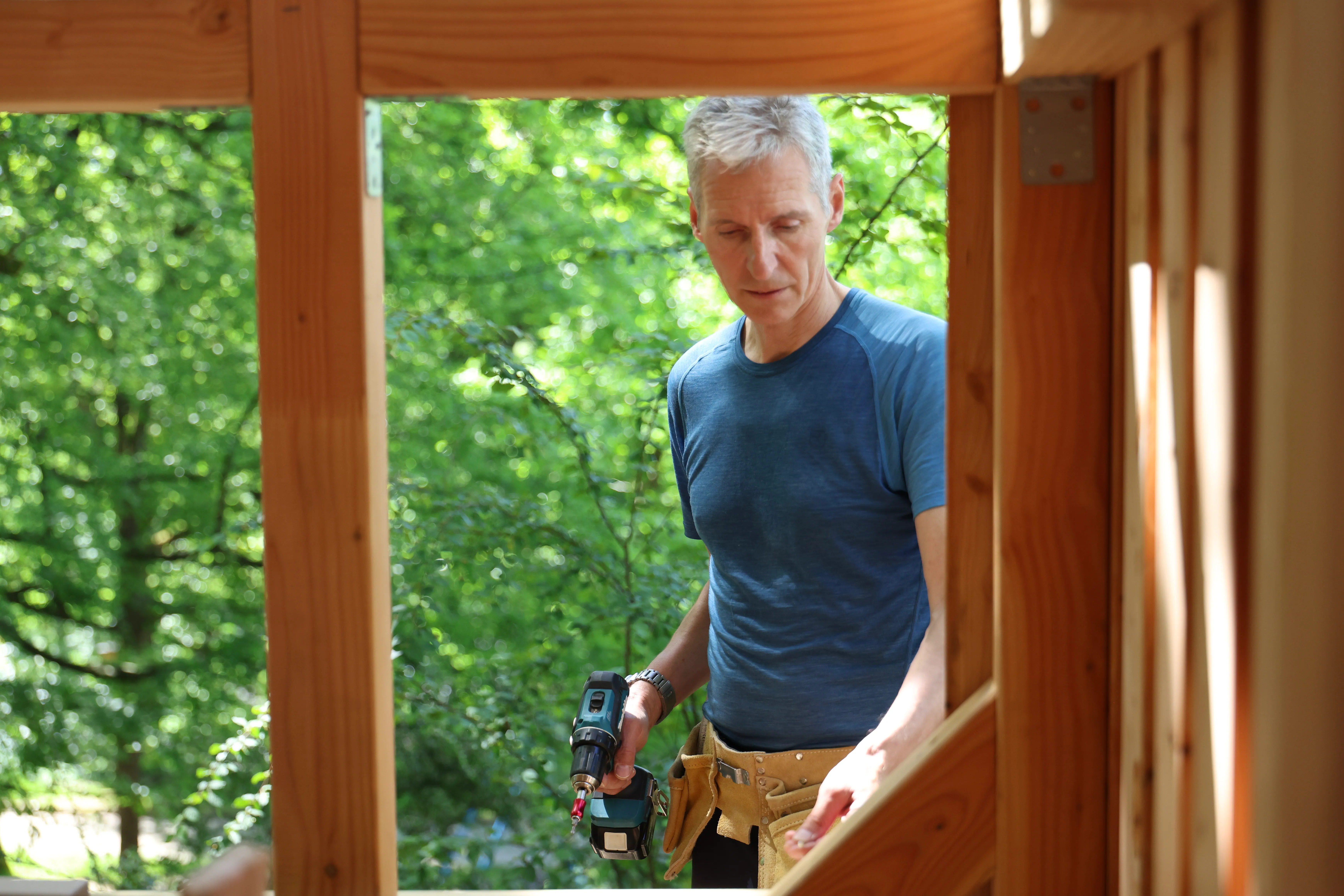 Ein Mann mittleren Alters mit grauem Haar schraubt an einem Holzbalken, trägt ein blaues T-Shirt und eine Werkzeugtasche.