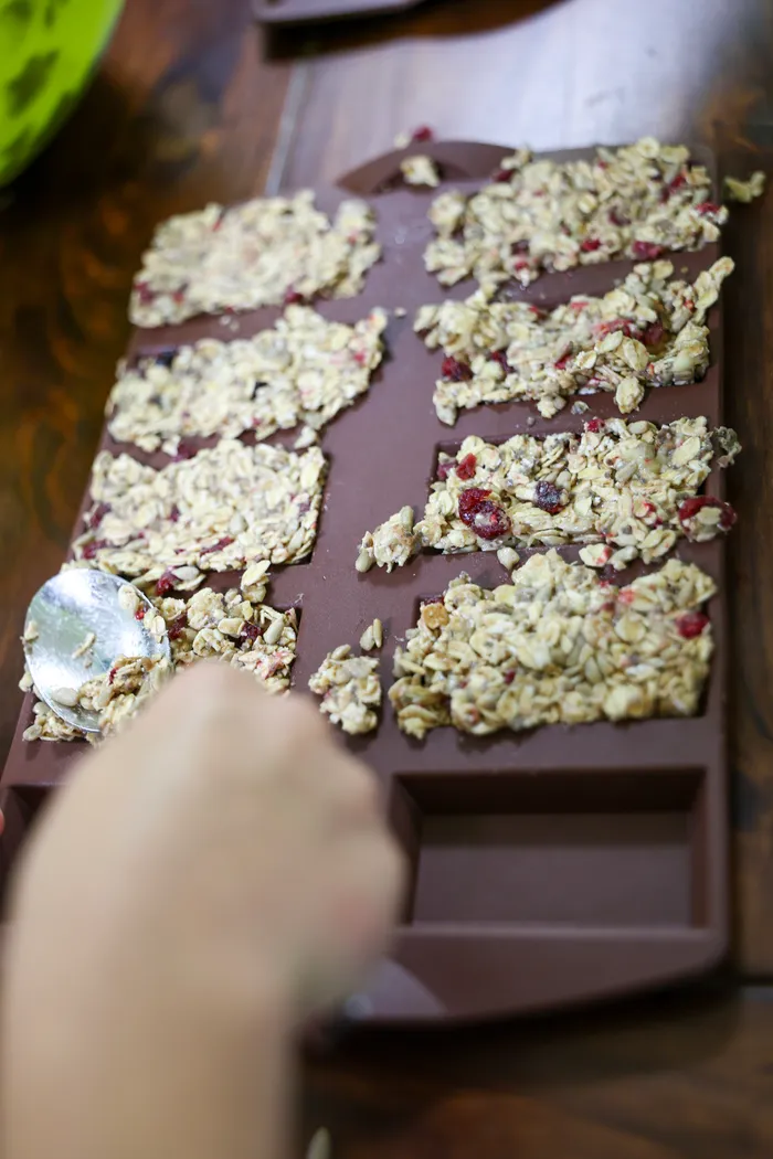 Ein Tablett mit kleinen, mit Haferflocken und getrockneten Früchten belegten Riegeln steht im Fokus. Eine Hand hält einen Löffel bereit, um die Riegel zu verteilen.