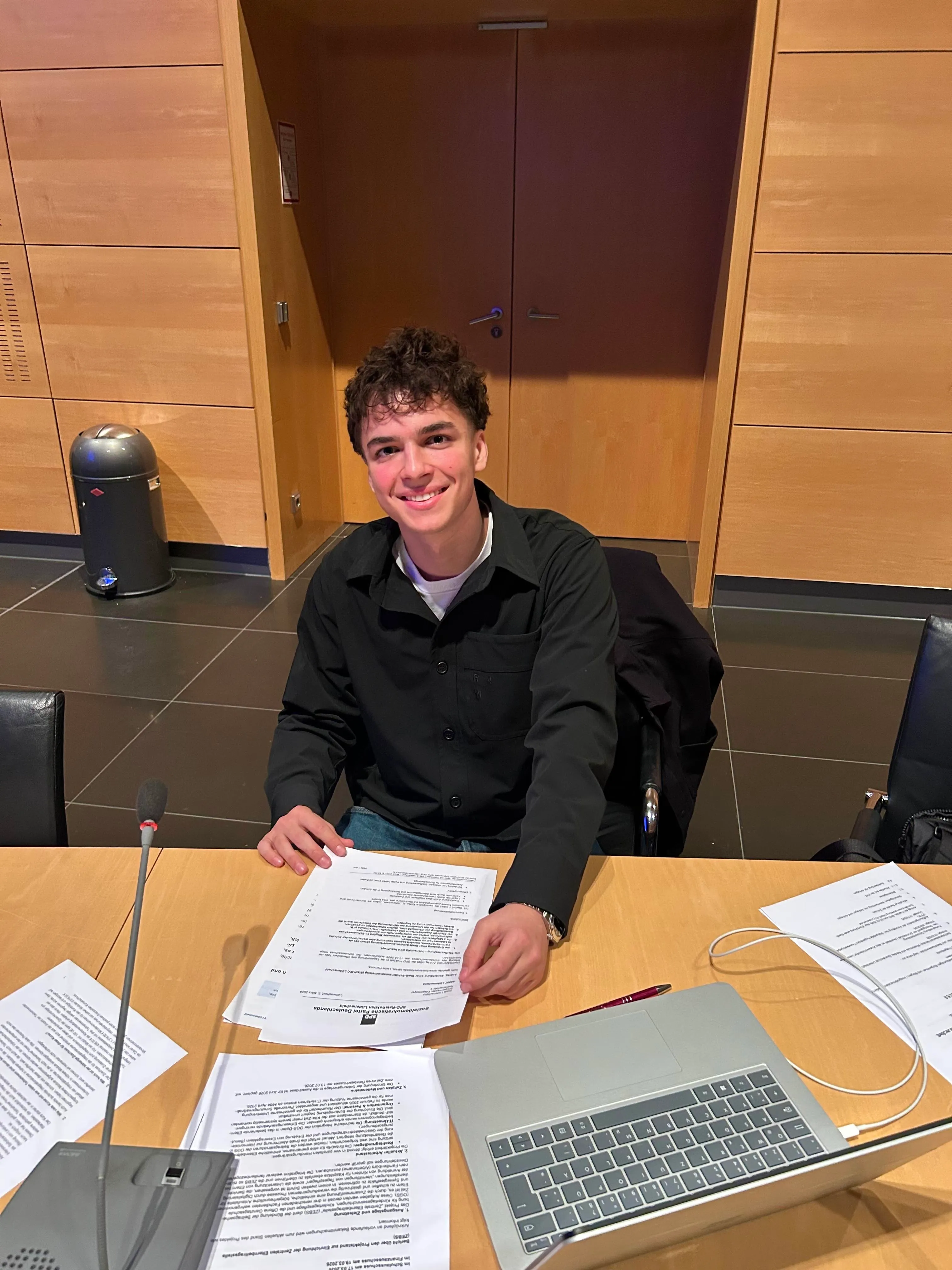 Ein junger Mann mit lockigem Haar lächelt in einem Konferenzraum, umgeben von Papieren und einem Laptop. Er sitzt an einem Tisch mit einem Mikrofon und unterzeichnet ein Dokument.