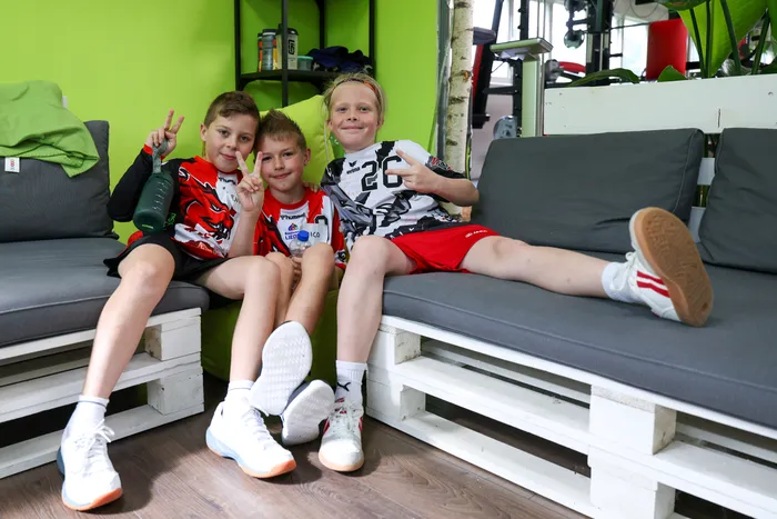 Drei Jungen sitzen lächelnd auf einer grauen Paletten-Couch. Sie tragen Sportkleidung und zeigen Victory-Zeichen. Im Hintergrund steht ein Regal mit Getränken und Snacks.