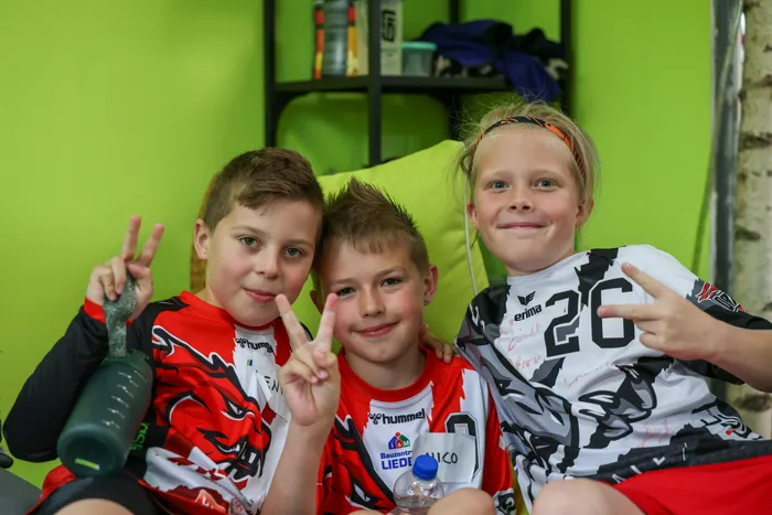 Drei Kinder im Alter von etwa 10 Jahren sitzen nebeneinander auf einer Couch und lächeln in die Kamera. Sie tragen rote und weiße Trikots mit verschiedenen Logos und geben mit ihren Händen das Victory-Zeichen. Im Hintergrund ist eine grüne Wand und Sportausrüstung zu sehen.