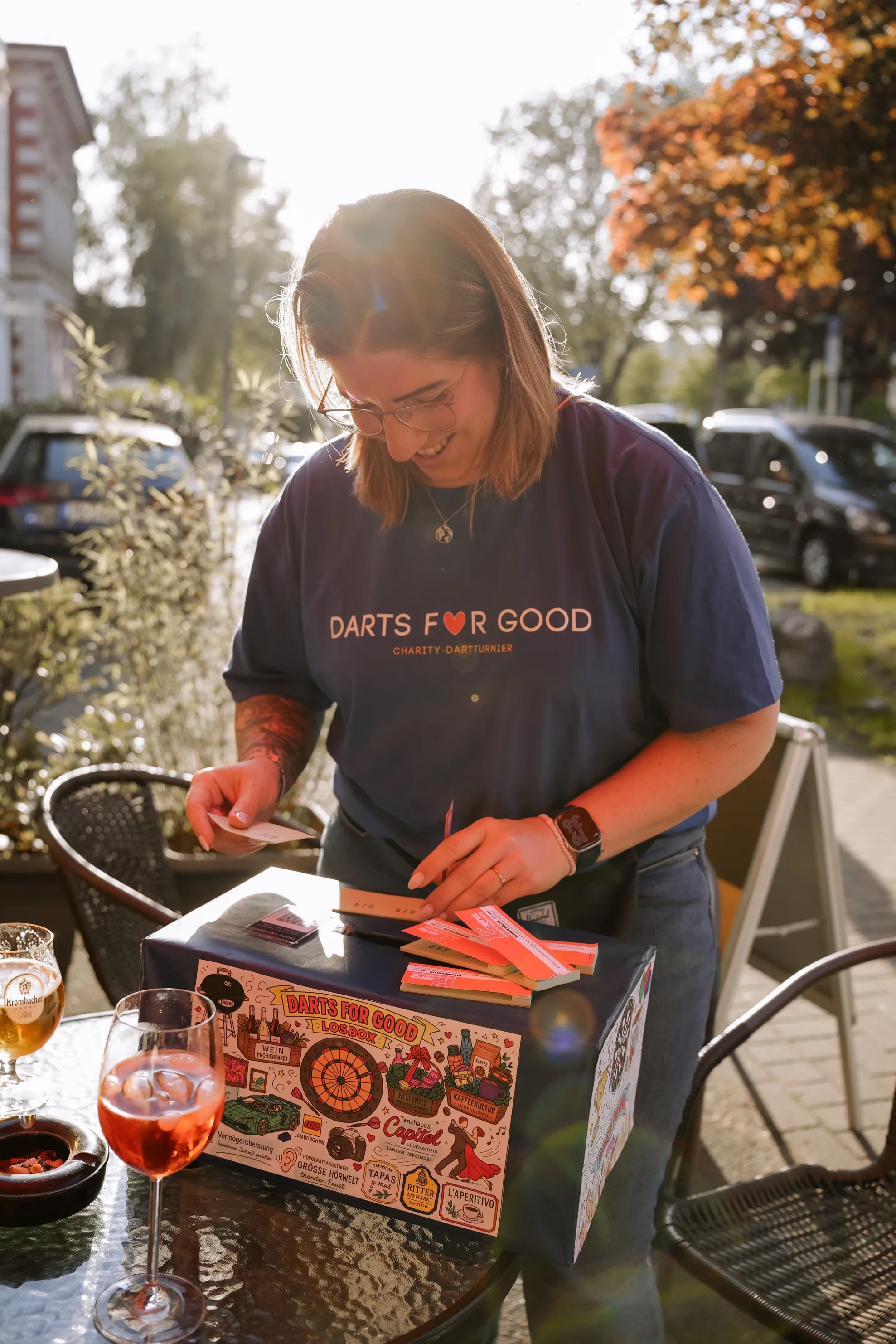 Eine Frau mit kurzem, blondem Haar und Brille packt ein buntes Dart-Set mit London-Motiven aus. Sie trägt ein T-Shirt mit der Aufschrift „Darts for Good – Charity Dartturnier“.