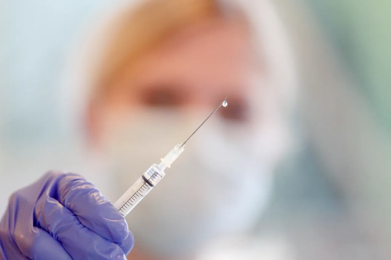 Eine Hand mit blauem Handschuh hält eine gefüllte Spritze, im Hintergrund verschwommen ein Arzt mit Mundschutz.