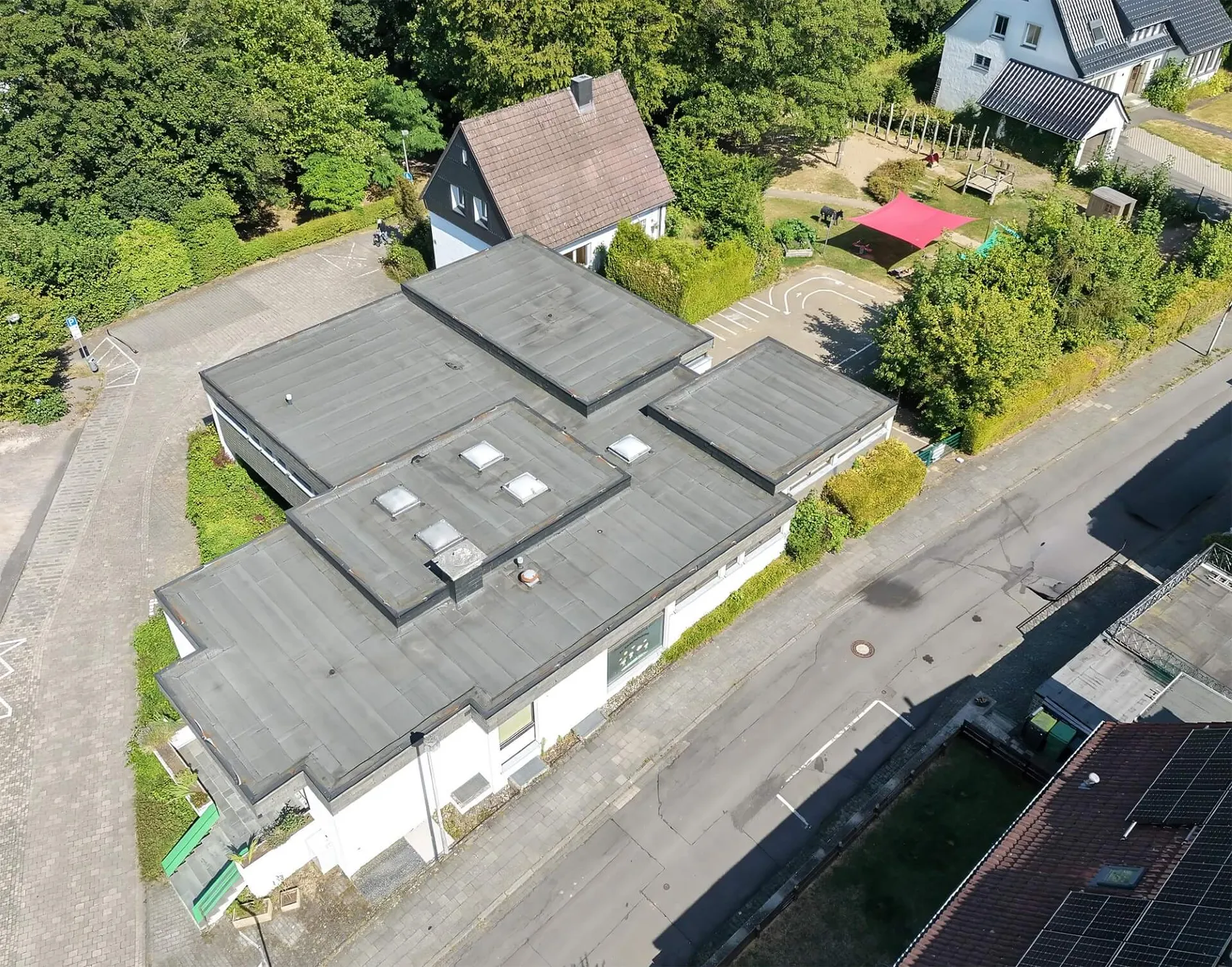 Luftaufnahme der Kindertageseinrichtung Hochstraße mit Flachdach, umgeben von Bäumen und einem Spielplatz mit rotem Sonnenschutz.