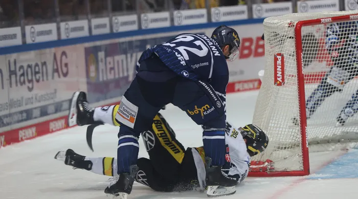 Eishockeyspieler Daniel Neumann in dunkelblauer Uniform ringt einen Gegenspieler in weiß-gelber Uniform vor dem Tor zu Boden.