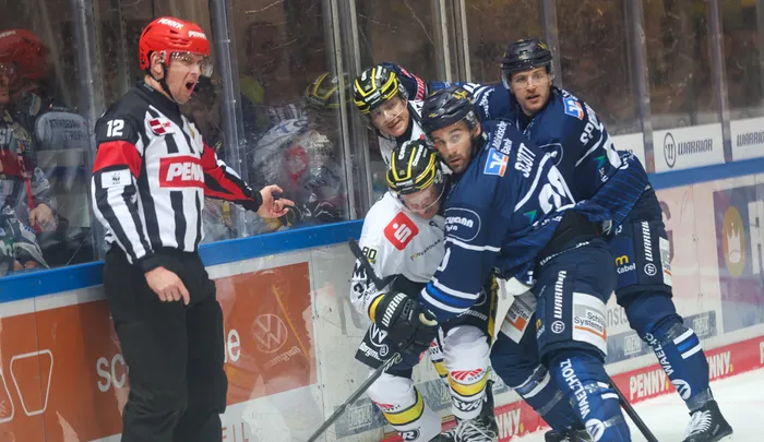 Ein Schiedsrichter in schwarz-weiß gestreiftem Trikot und rotem Helm schreit während eines Eishockeyspiels auf Spieler in blauen und weißen Trikots ein.