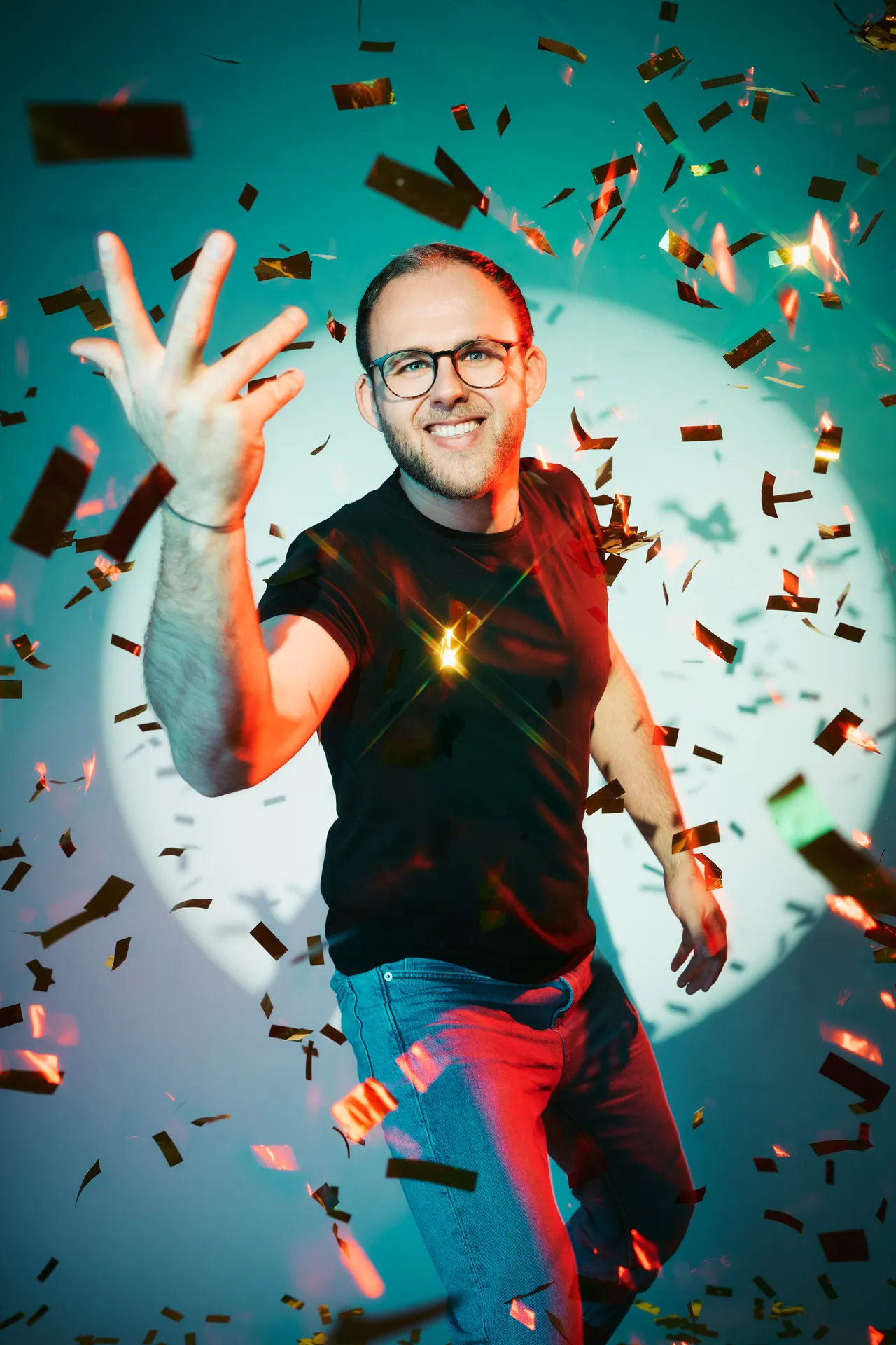Mann mit Brille und schwarzem T-Shirt steht vor einem bunten Lichtkreis und streckt eine Hand in die Luft, umgeben von Konfetti.