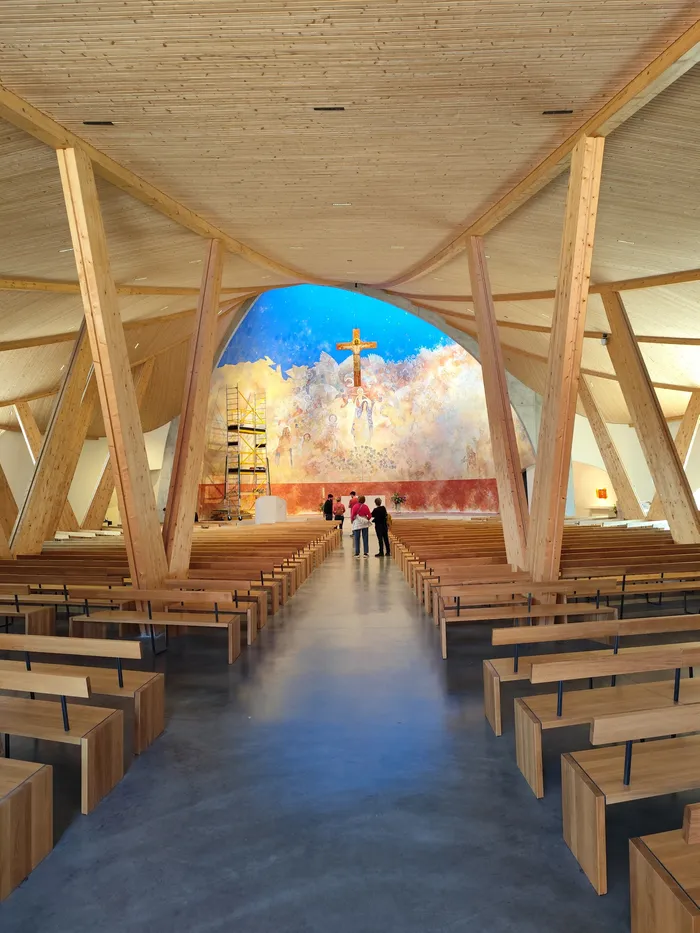 Innenraum einer hellen Kirche mit Holzbänken und einem großen Wandgemälde mit Kruzifix, vor dem sich einige Personen aufhalten. Das Dach besteht aus hellen Holzkonstruktionen.