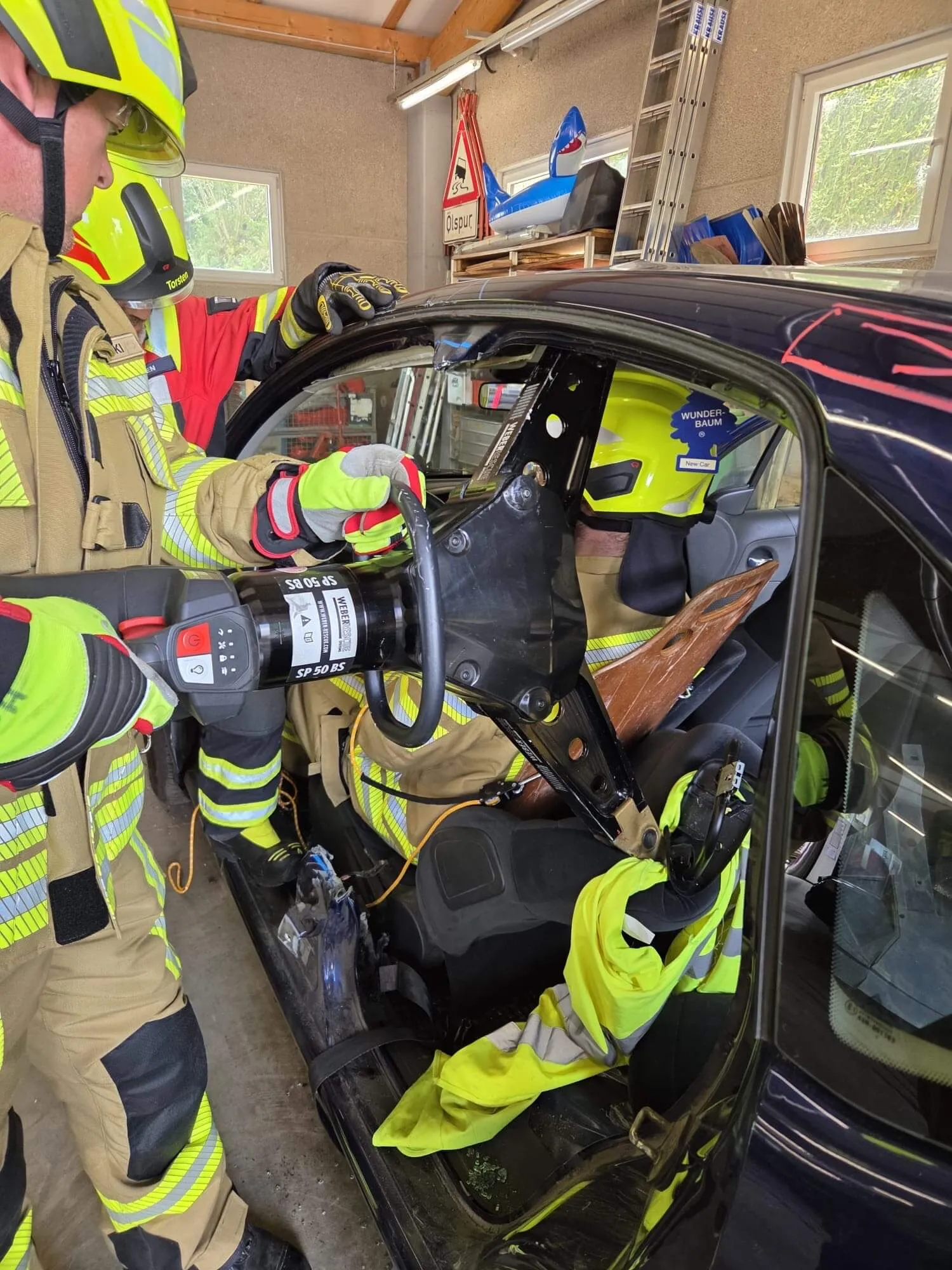 Zwei Feuerwehrleute in voller Schutzkleidung befreien eine Person aus einem stark beschädigten Auto. Im Hintergrund sind weitere Einsatzkräfte und Rettungsequipment zu sehen.