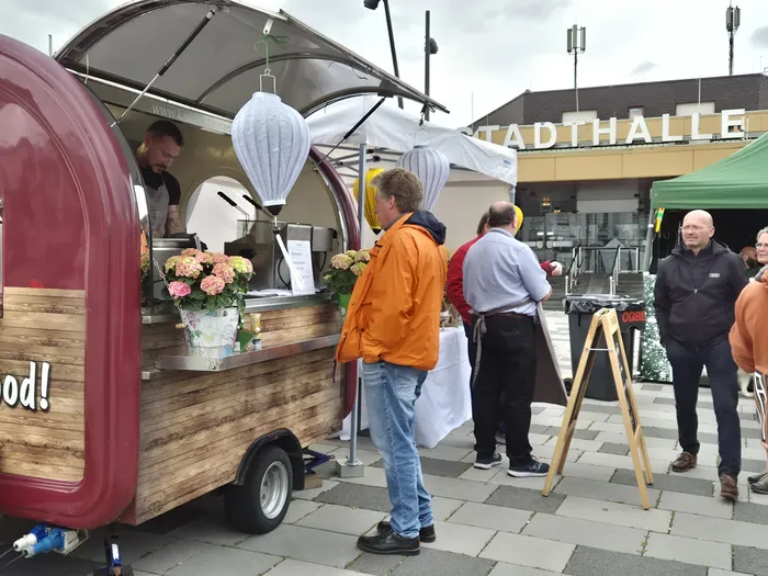 Ein rot-brauner Imbisswagen mit Blumen vor dem Eingang steht auf einem Platz vor der Stadthalle Meinerzhagen. Mehrere Personen stehen vor dem Wagen und unterhalten sich. Ein Mann mit Tattoos bedient im Wagen.