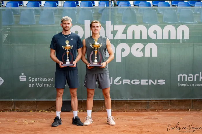 Zwei Männer halten Goldtrophäen vor einer Platzmann Open-Werbewand.