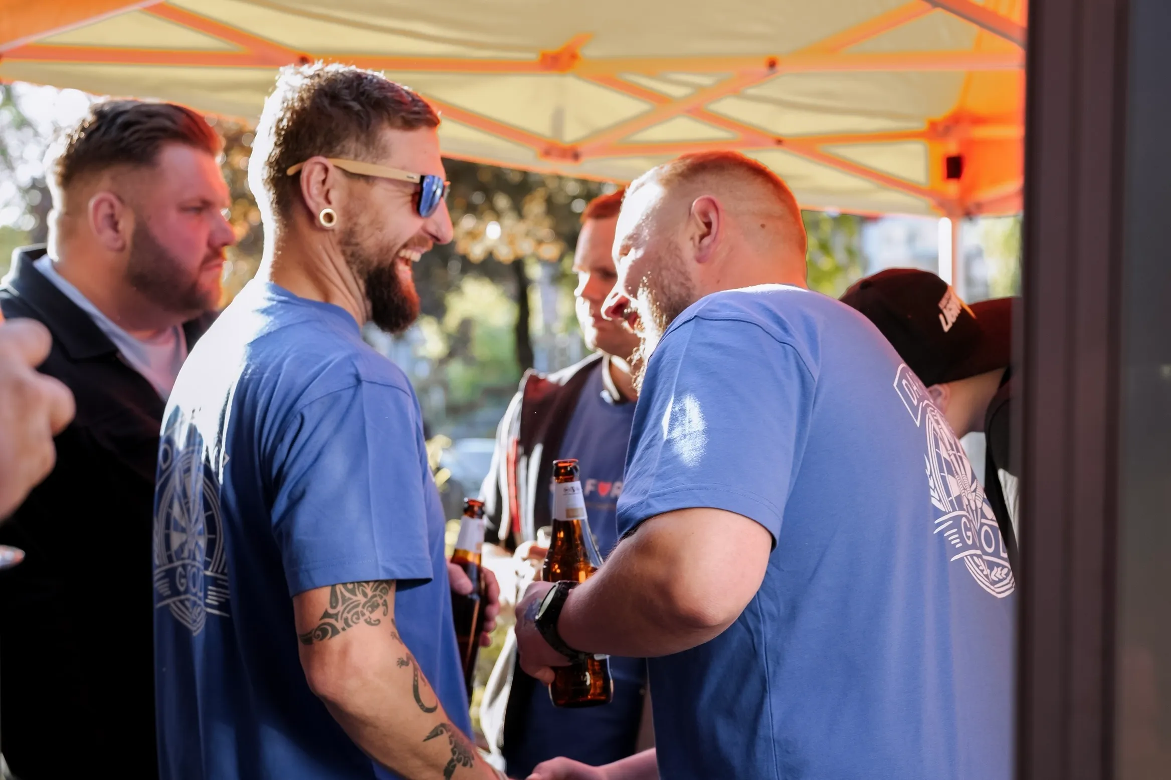 Drei Männer umarmen sich lachend im Freien, unter einem orangefarbenen Sonnenschutz. Sie tragen blaue T-Shirts und Sonnenbrillen.