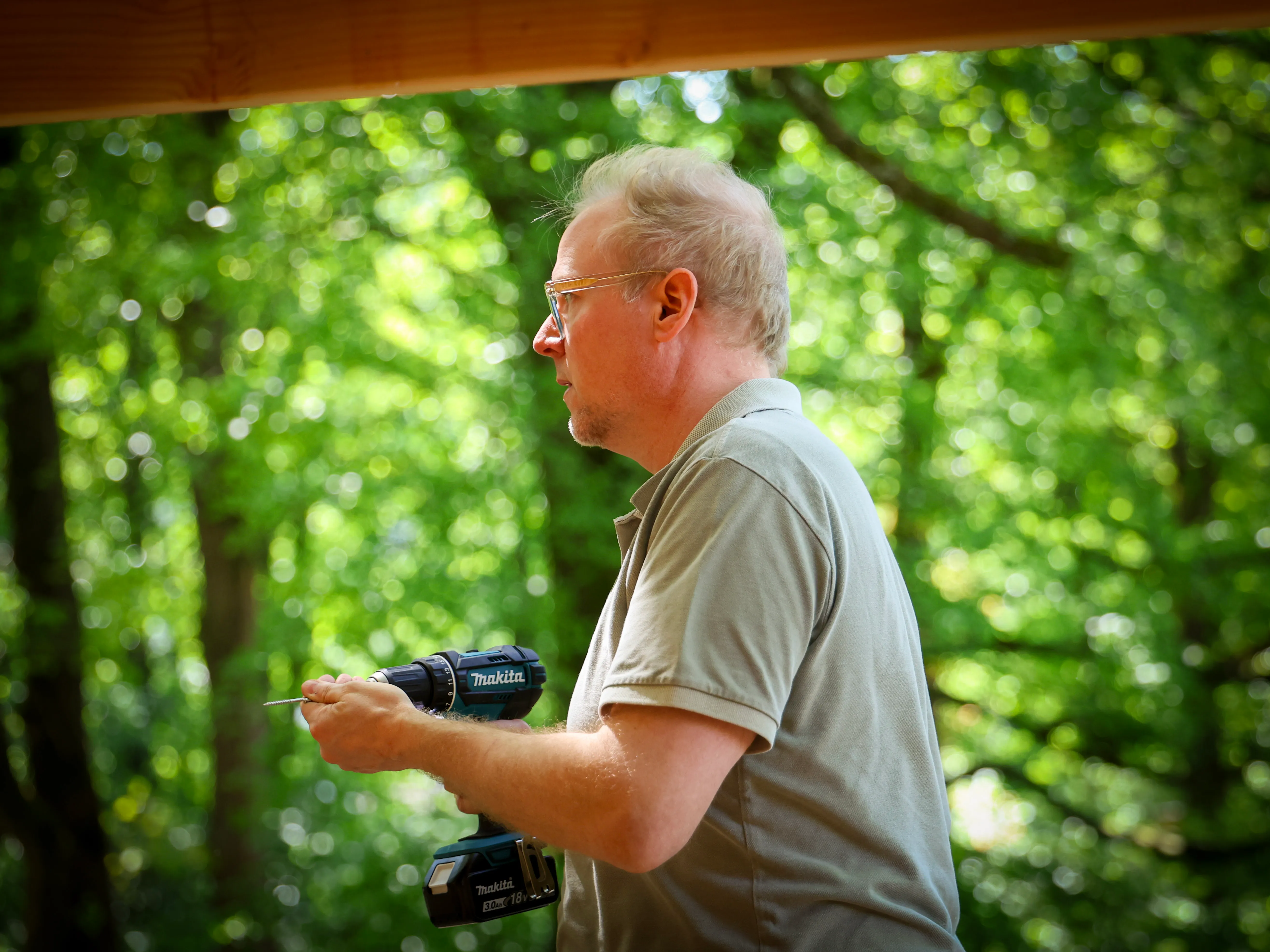 Ein Mann mit Brille und T-Shirt hält einen Akkuschrauber in der Hand und arbeitet an einem Holzbauwerk im Freien. Im Hintergrund ist dichtes Grün zu sehen.
