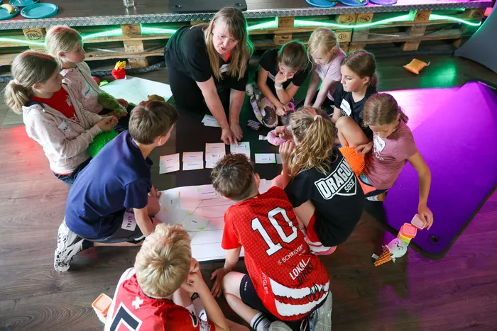 Eine Frau erklärt einer Gruppe von Kindern im Kreis liegend das Thema gesunde Ernährung anhand von Bildkarten. Einige Kinder tragen Sportkleidung.