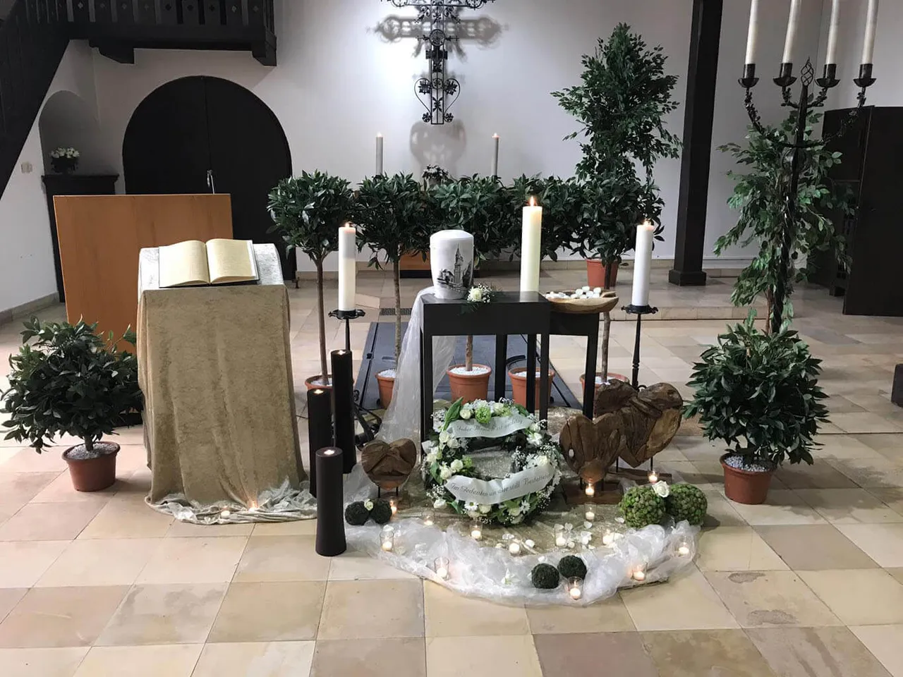 Gedenkarrangement in einer Kapelle mit einem aufgeschlagenen Buch, großen weißen Kerzen, einem Blumenkranz und hölzernen Herzen auf dem Boden.