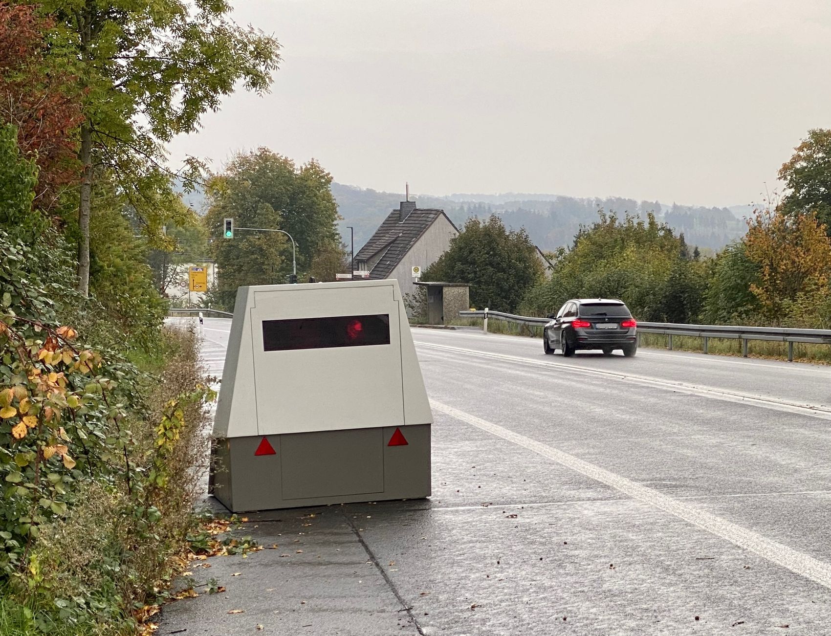 Ein grauer Blitzeranhänger steht am Straßenrand, während ein schwarzes Auto auf der Straße vorbeifährt. Im Hintergrund sind Bäume und ein Gebäude zu sehen.