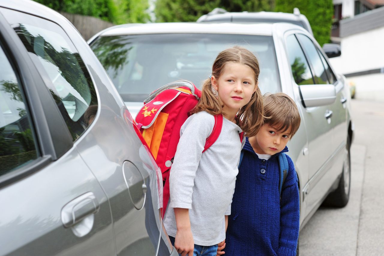 Zwei Kinder mit Rucksäcken stehen zwischen zwei geparkten Autos auf einer Straße.