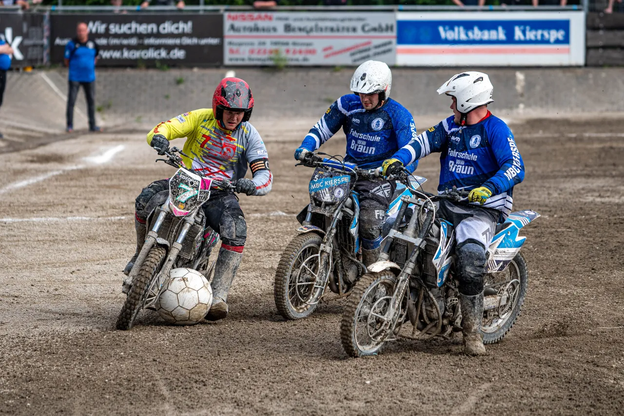 Drei Motorradfahrer spielen auf einer staubigen Fläche einen Fußball. Die Fahrer tragen Helme und Schutzkleidung in verschiedenen Farben und steuern ihre Motorräder, um den Ball zu bewegen. Im Hintergrund sind Zuschauer und Werbebanner zu sehen.