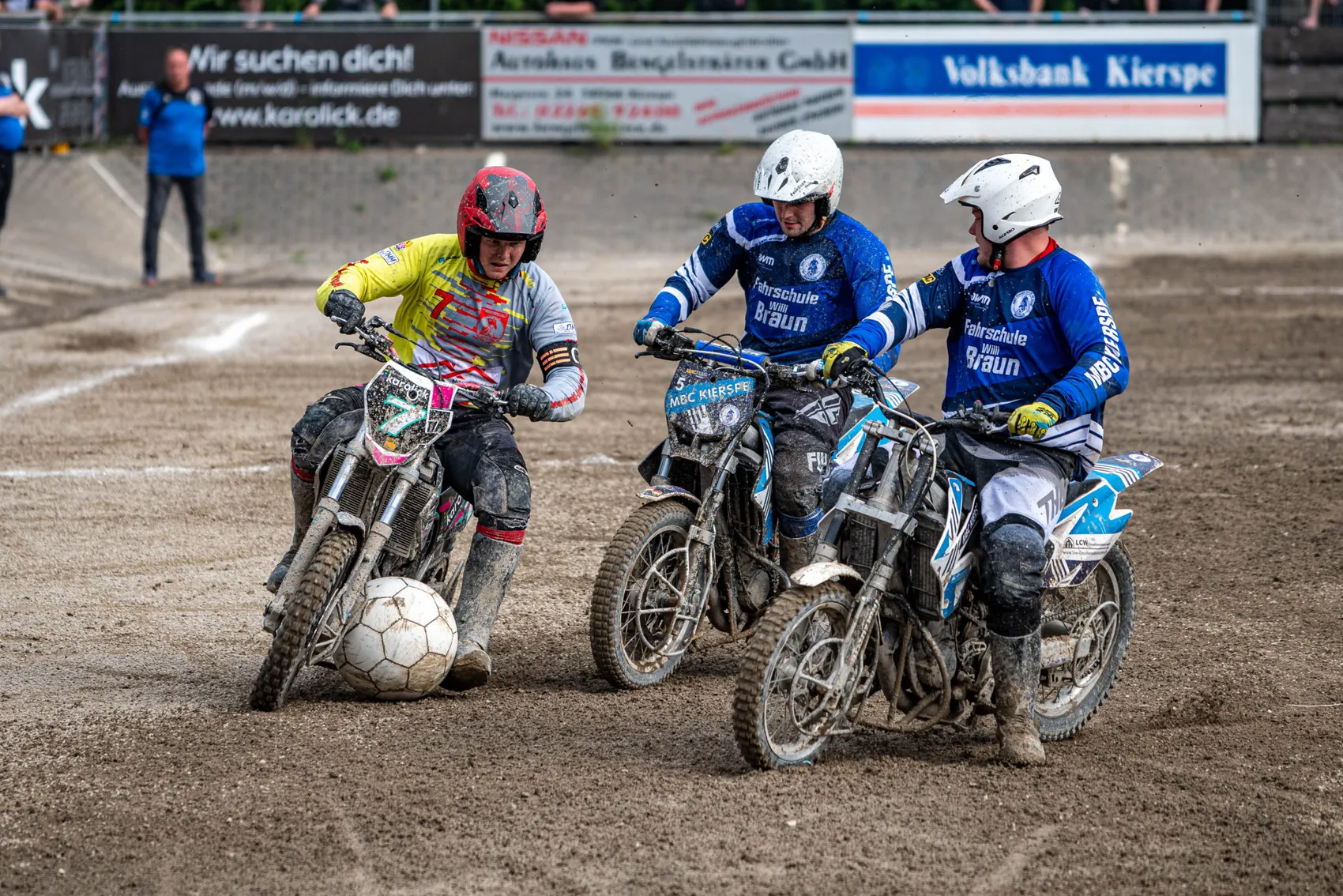 Drei Motorradfahrer spielen auf einer staubigen Fläche einen Fußball. Die Fahrer tragen Helme und Schutzkleidung in verschiedenen Farben und steuern ihre Motorräder, um den Ball zu bewegen. Im Hintergrund sind Zuschauer und Werbebanner zu sehen.