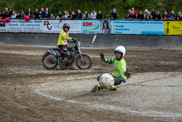 Ein Motoballspieler in Schutzkleidung und Helm versucht, einen großen Ball auf einer staubigen Strecke zu kontrollieren, während ein Motorradfahrer im Hintergrund fährt. Zuschauer säumen die Strecke.