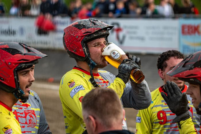 Ein Motoballspieler in rotem Helm trinkt aus einer Bierflasche, umgeben von Teamkollegen in gelben Trikots. Im Hintergrund sind weitere Spieler und Zuschauer zu sehen.