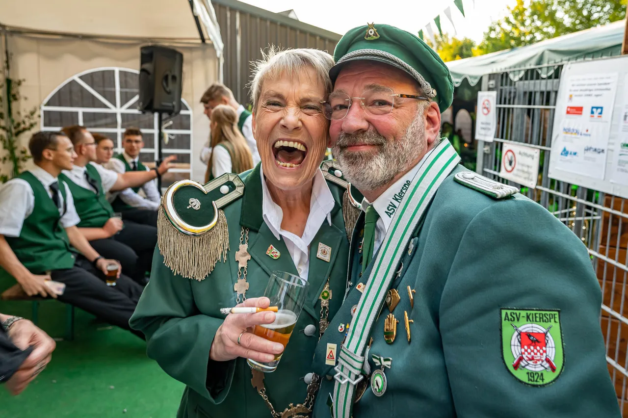 Ilkra Krause und Holger Lüddemann in grünen Schützenuniformen lachen gemeinsam bei einem Schützenfest. Die Frau hält ein Glas Bier und eine Zigarette.