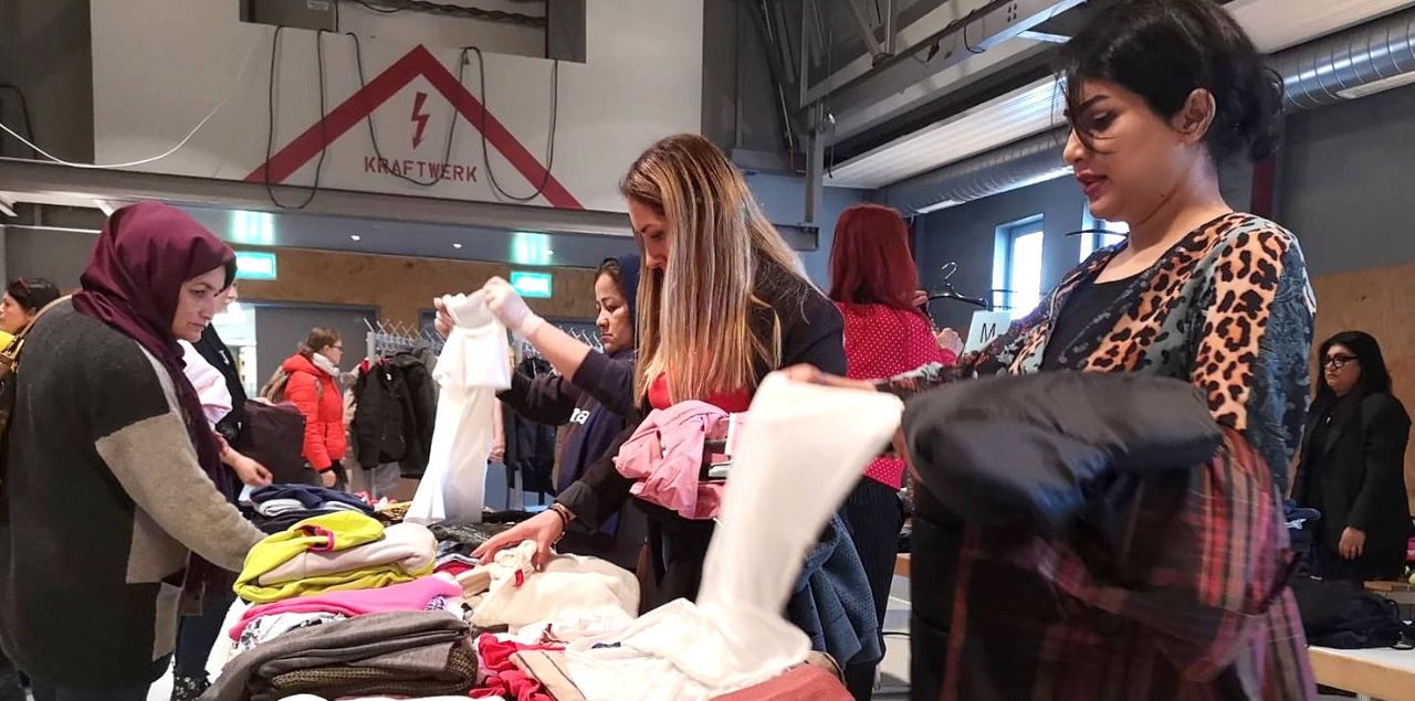 Mehrere Frauen wählen Kleidung auf einem Tauschmarkt in einer Turnhalle aus. Im Hintergrund ein Banner mit dem Schriftzug „Kraftwerk“.