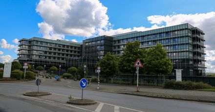 Das Gebäude des Klinikums Lüdenscheid mit moderner Glasfassade und umliegender Bepflanzung. Im Vordergrund sind Verkehrsschilder und eine Straße zu sehen.