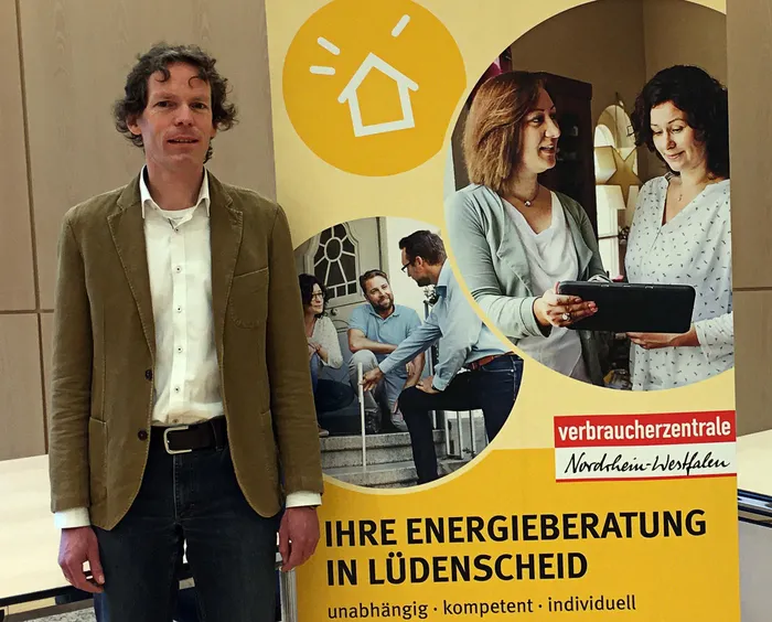 Ein Mann mit lockigem Haar im beigen Sakko steht vor einem Werbeplakat für Energieberatung der Verbraucherzentrale Nordrhein-Westfalen in Lüdenscheid. Das Plakat zeigt Menschen, die sich über ein Tablet informieren.
